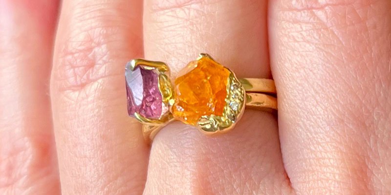 Two-stone ring with rough purple and orange gemstones on a gold band, worn on a finger.