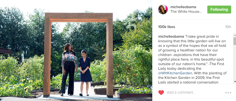 Michelle Obama and a young girl standing in a garden under a wooden archway.