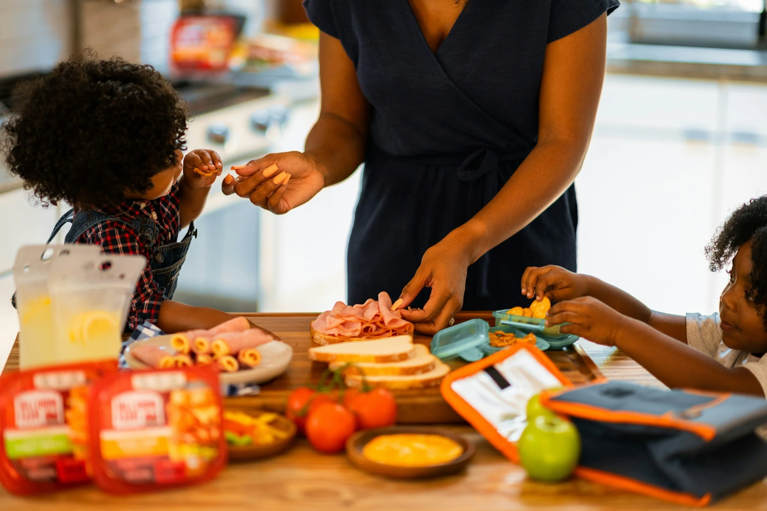 a woman and children prepare meals in the kitchen while snacking at the kitchen island.