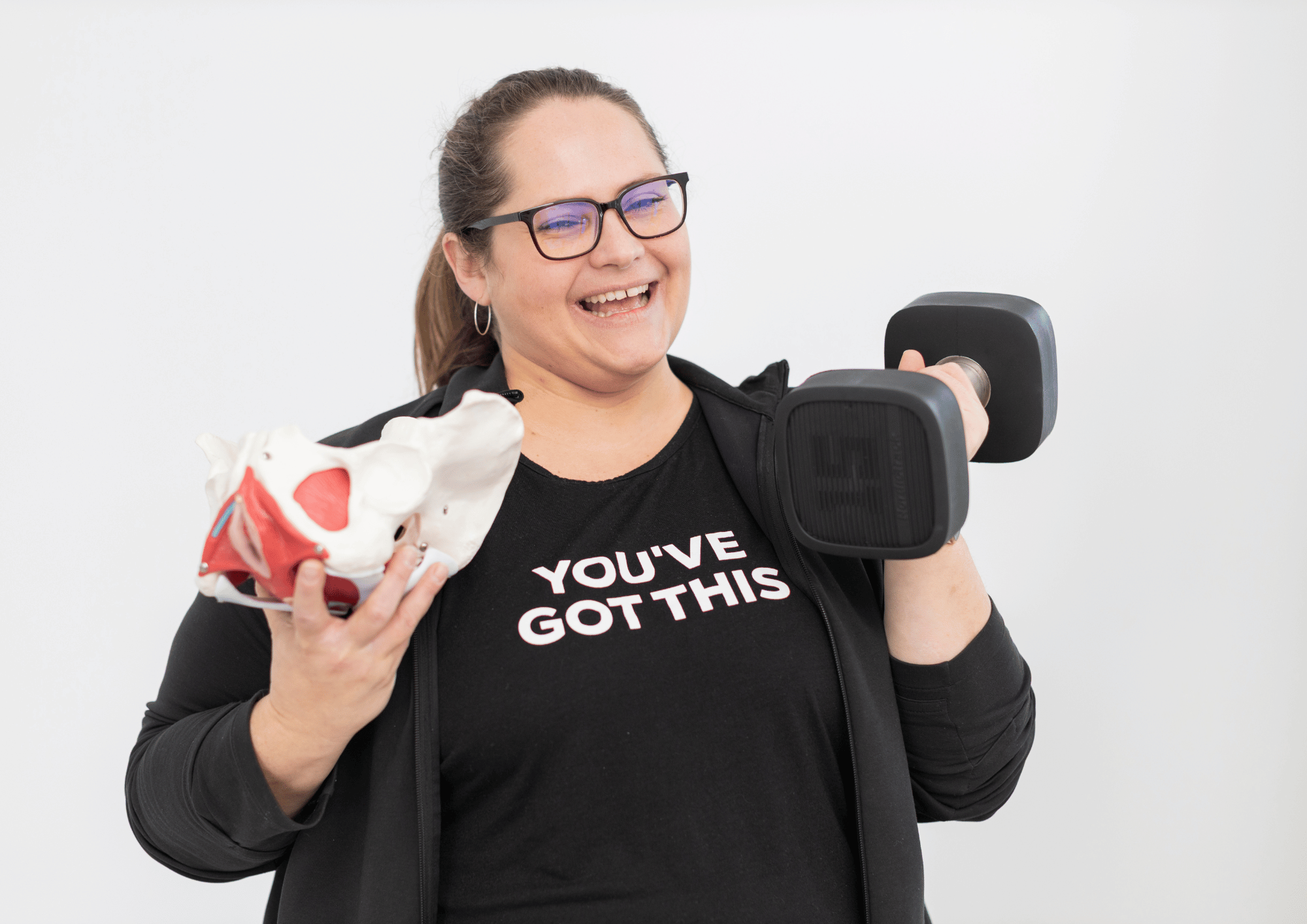 A woman with long brown hair wearing a shirt that says, you've got this. Holding a 15 lb. weight and a plastic pelvis.