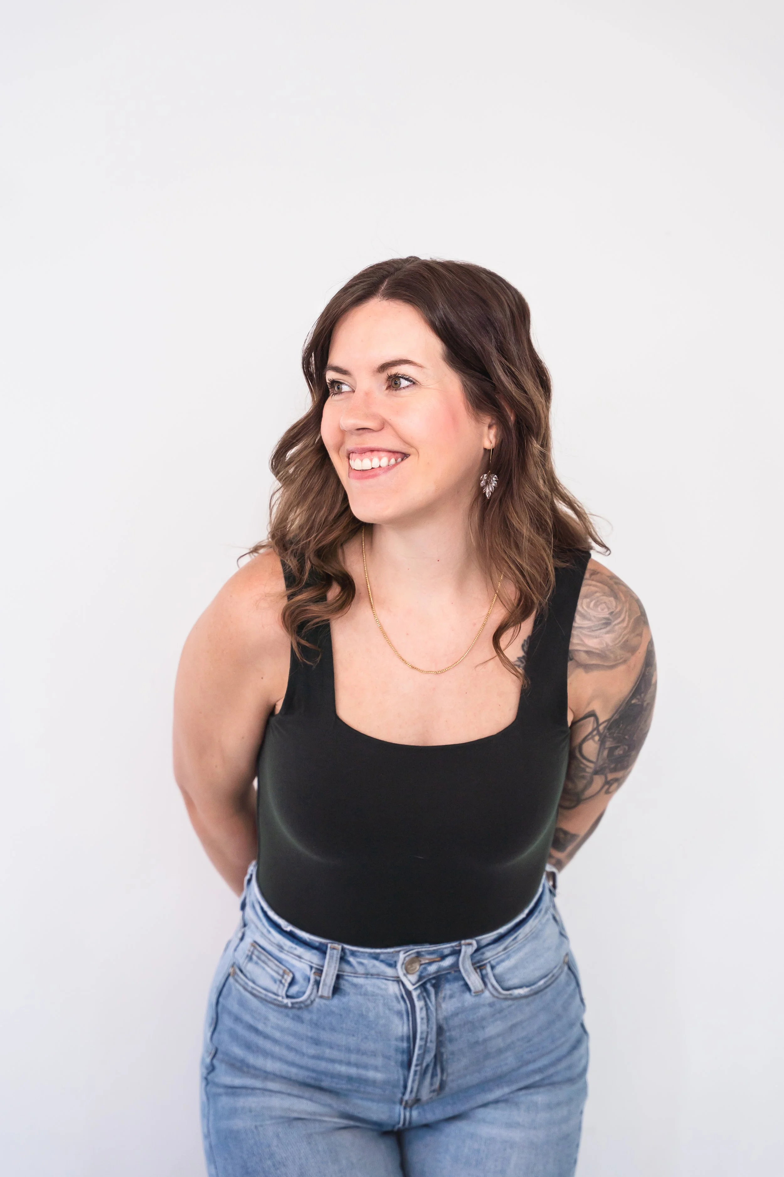 A smiling woman with shoulder-length wavy brown hair, wearing a black sleeveless top, light blue jeans, gold necklace, and earrings, standing against a plain white background.