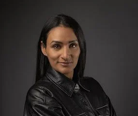 A woman with long dark hair and a black leather jacket, looking confidently into the camera, against a dark background.