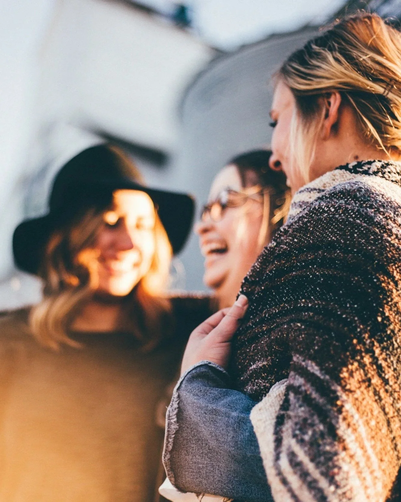 Three women laughing and smiling together, engaging in conversation outdoors during sunset.