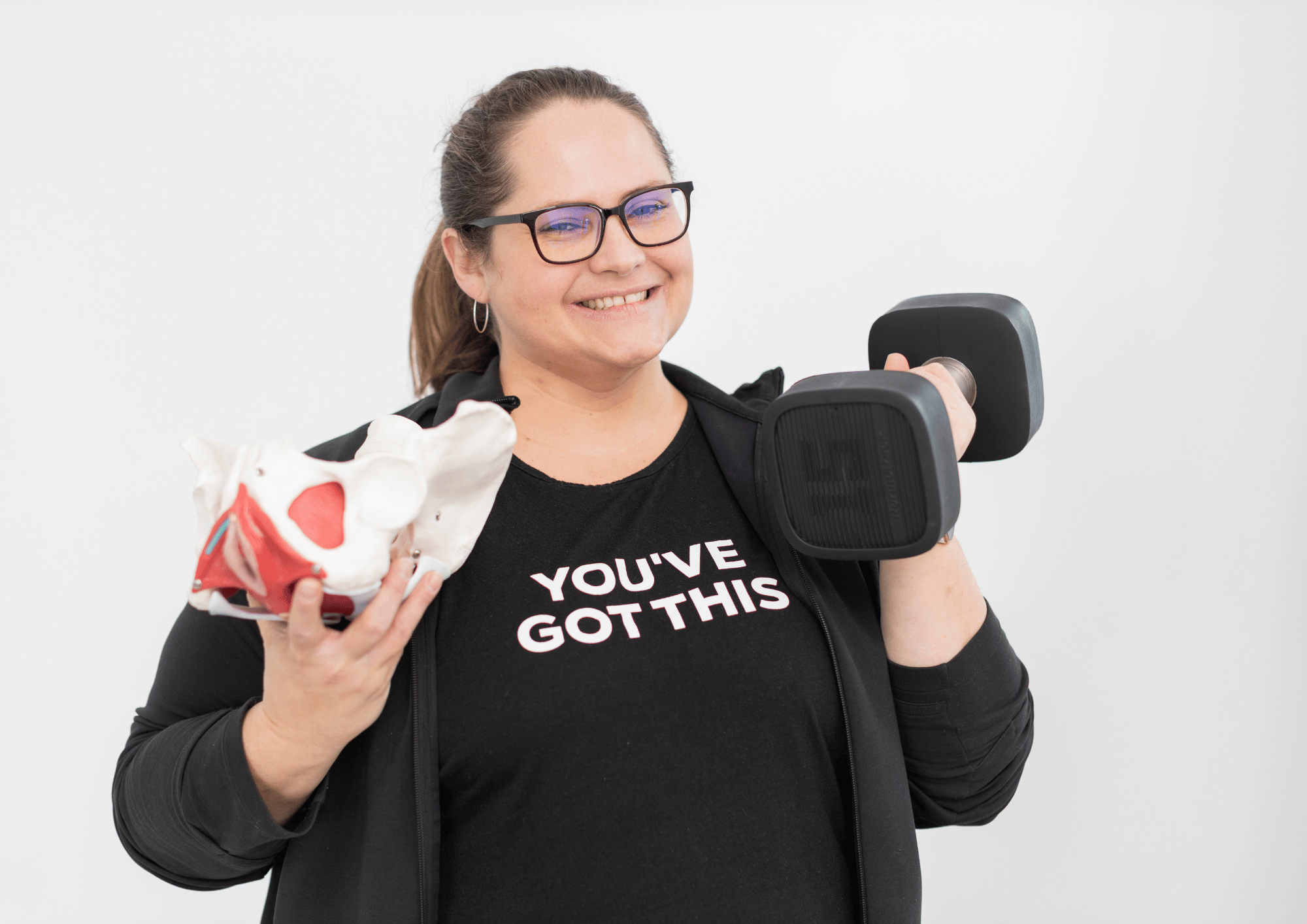 a happy woman holds a 15 lb. weight and a plastic pelvis with a shirt that says, you've got this