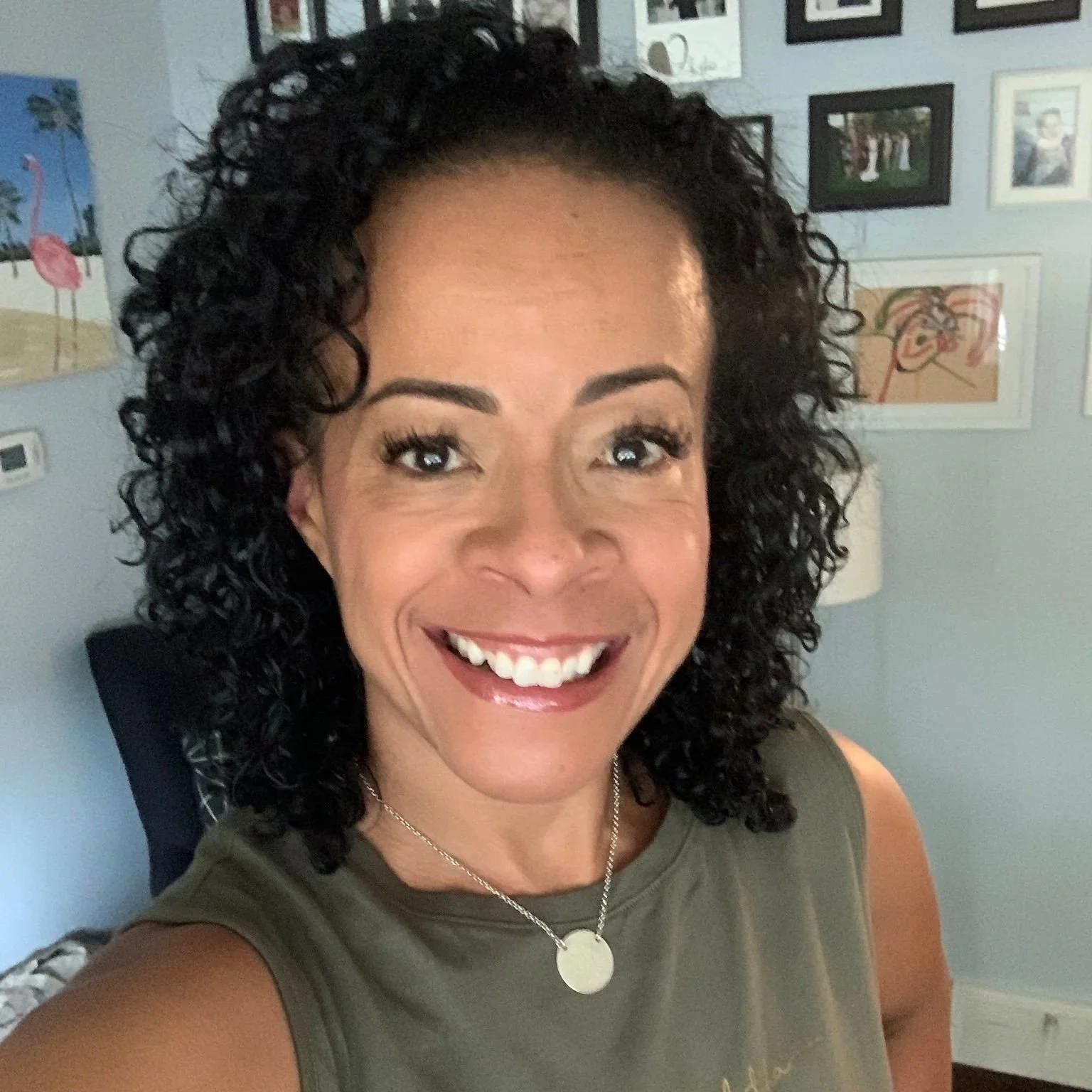 A woman with dark, curly hair and bright smile taking a selfie in a room with framed artwork hanging on the wall behind her.