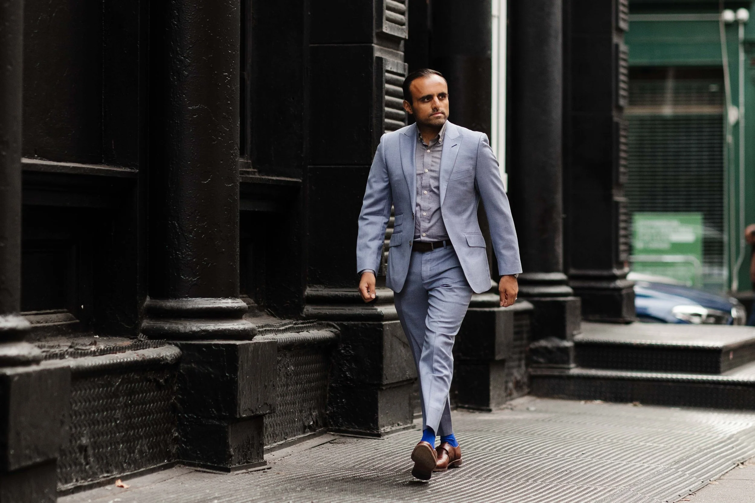 A man in a light blue suit walking on a city sidewalk, with tall dark buildings and a glass building in the background.