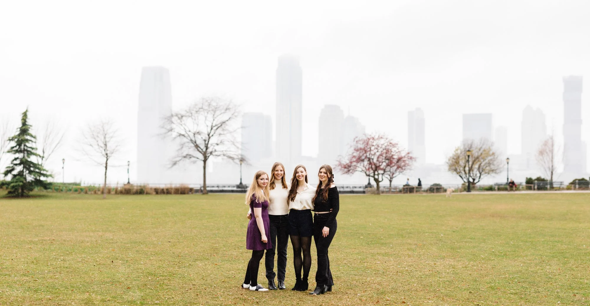 Four women standing in a park with a city skyline and bare trees in the background.