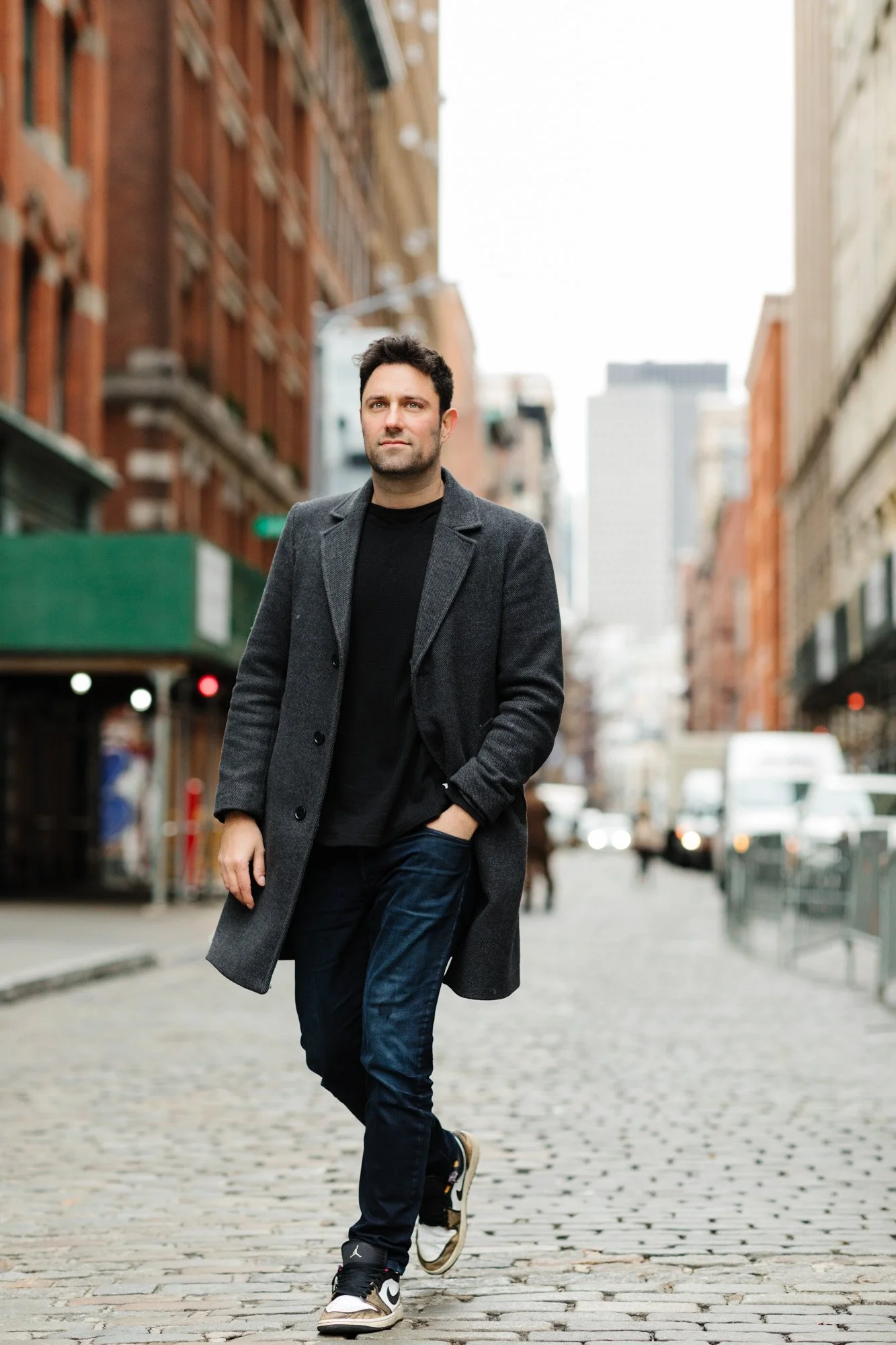 Man walking on a cobblestone street in a city, wearing a gray coat, black shirt, blue jeans, and sneakers.