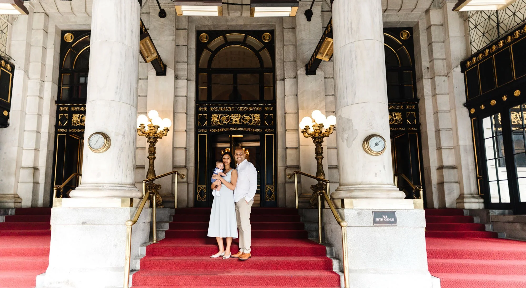 Family standing on red carpeted steps in front of a grand building entrance with large columns and decorative lamps.