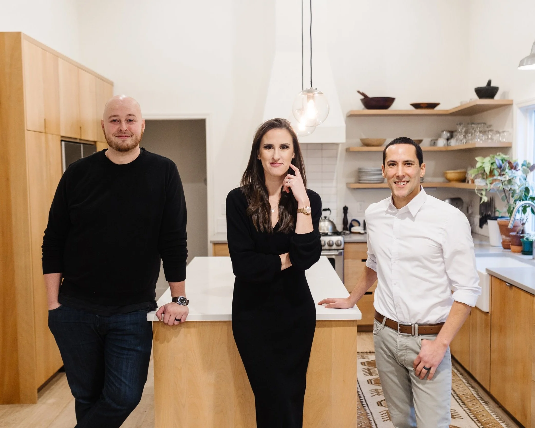 Three people in a modern kitchen with light wood cabinets, standing around a kitchen island, with plants on the counter and open shelving in the background.