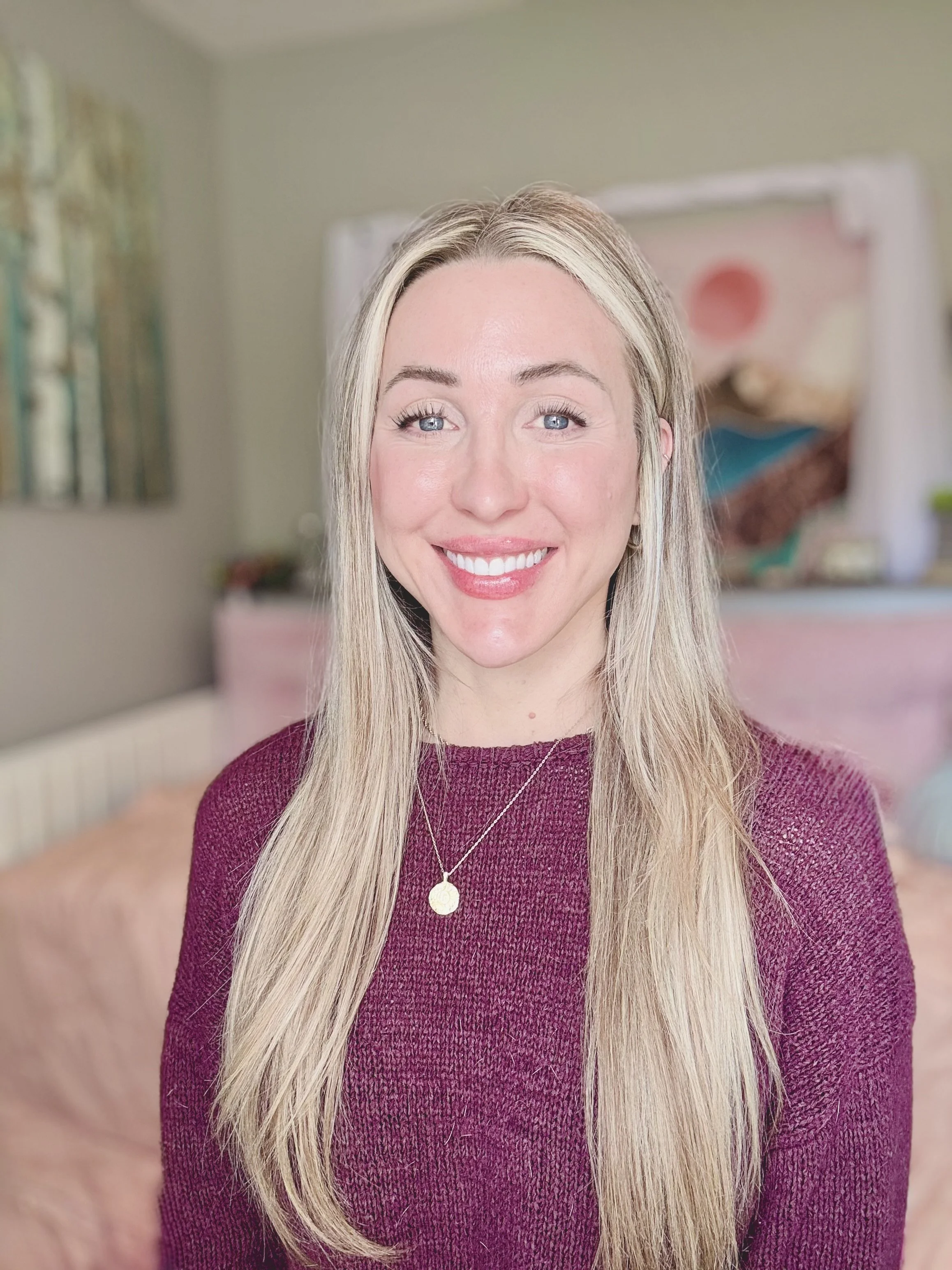 Portrait of a smiling woman with long blonde hair, wearing a maroon sweater and a silver necklace, inside a room with colorful artwork and a blurred background.