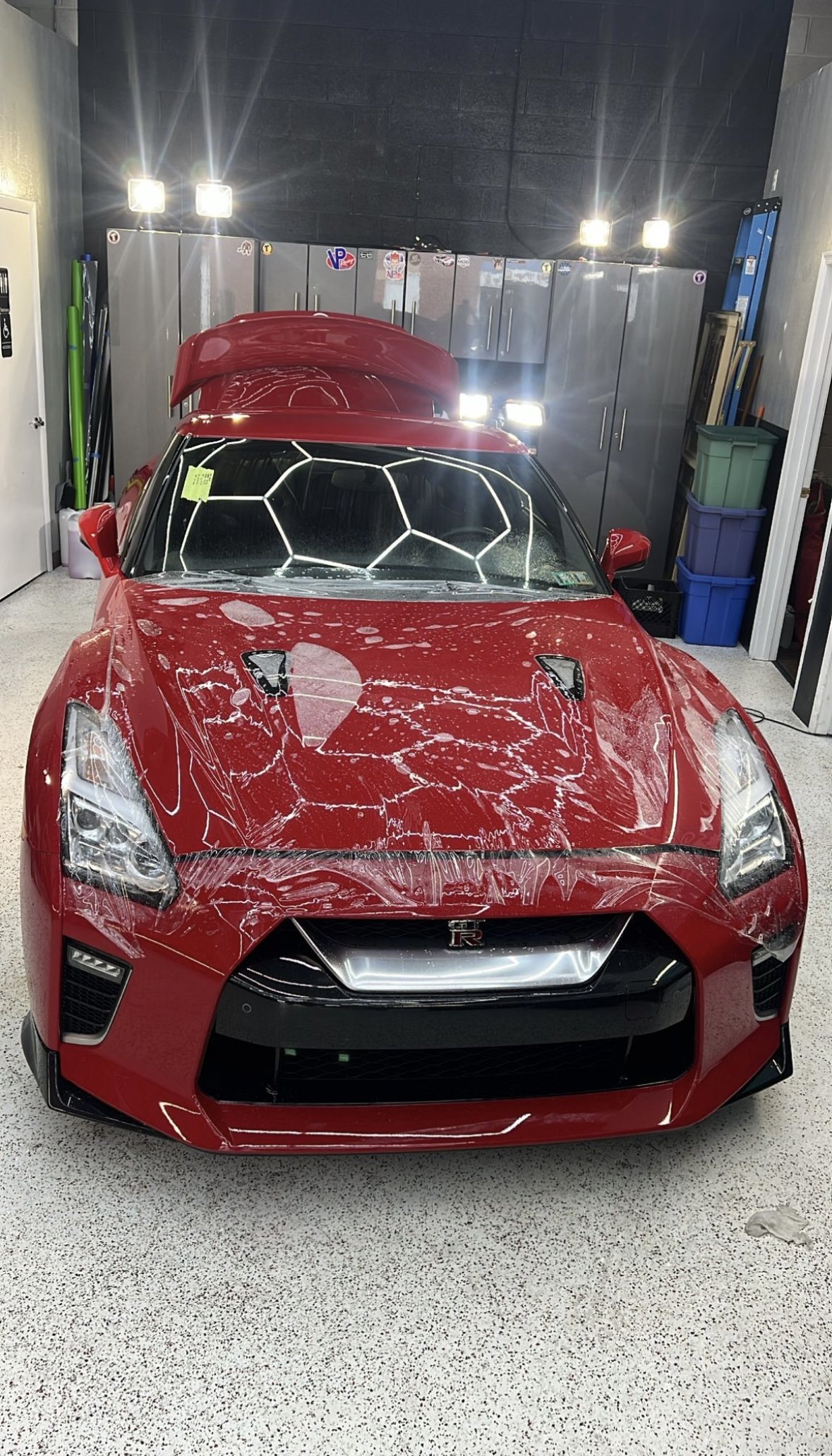 A red Nissan GT-R sports car in a garage with a damaged hood, windshield covered in soap suds, and plastic wrap on the front bumper. The garage has gray cabinets, storage bins, and bright overhead lighting.