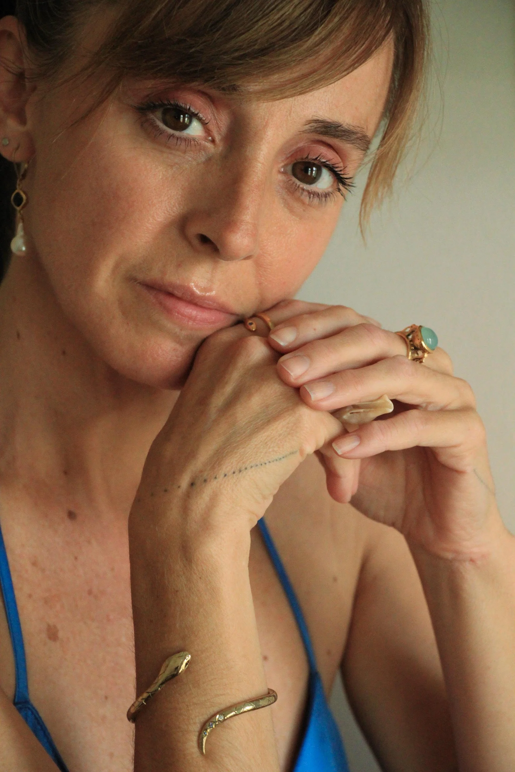 Close-up of a woman with light brown hair, brown eyes, and light skin, wearing gold jewelry and a blue top. She is resting her hands near her face.