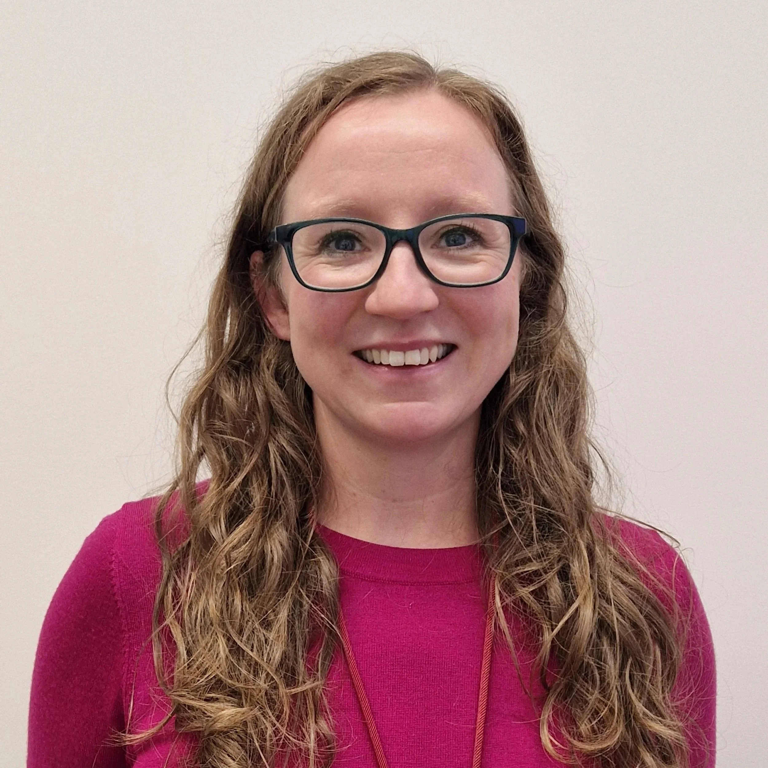 Katie is a white woman with long light brunette hair, she wears black glasses and a pink jumper, smiling to the camera.