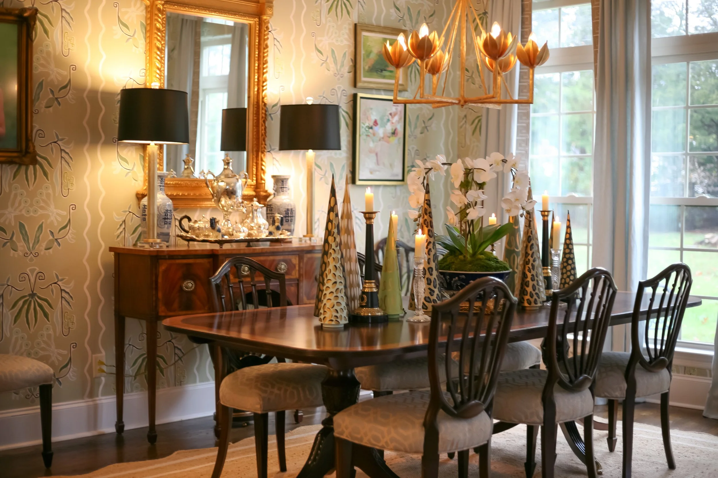 A dining room with a wooden table and six upholstered chairs, decorated with candles and a plant centerpiece. A sideboard with vases, lamps, and a large mirror hanging above it. The room has floral wallpaper, framed paintings, and large windows with 