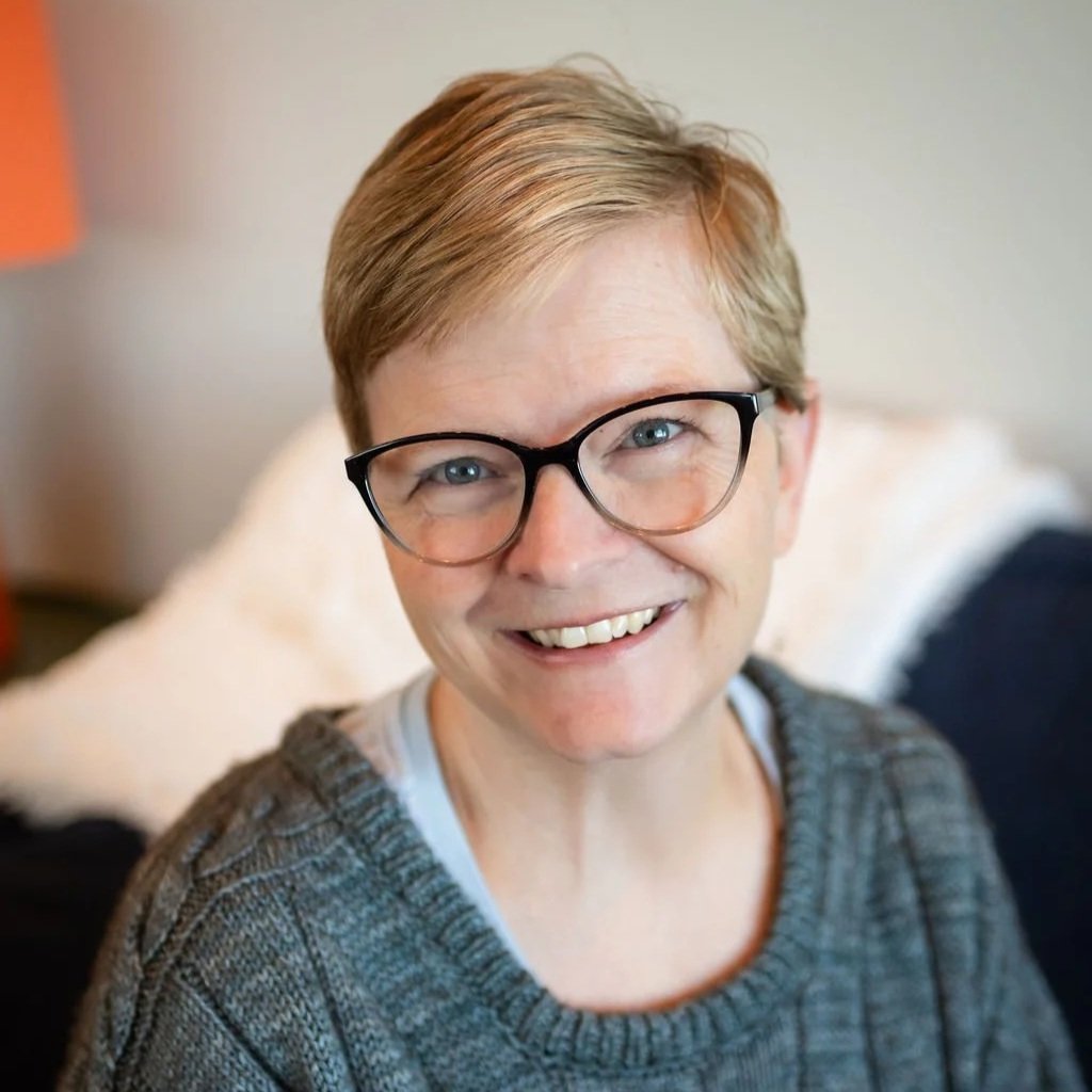 A smiling woman with glasses and short blonde hair, wearing a dark gray CB&W Colorado Birth & Wellness t-shirt, standing in front of a blue wall with a textured hanging art piece made of hanging threads.