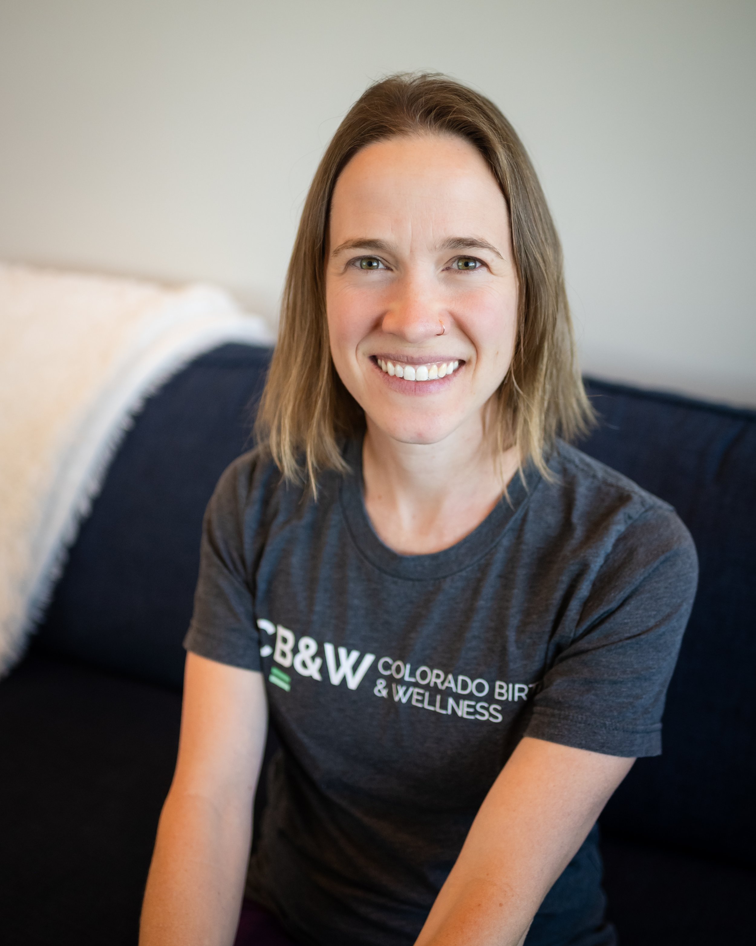 A smiling woman with shoulder-length brown hair and a septum piercing standing in front of a teal wall with a decorative yarn art piece.