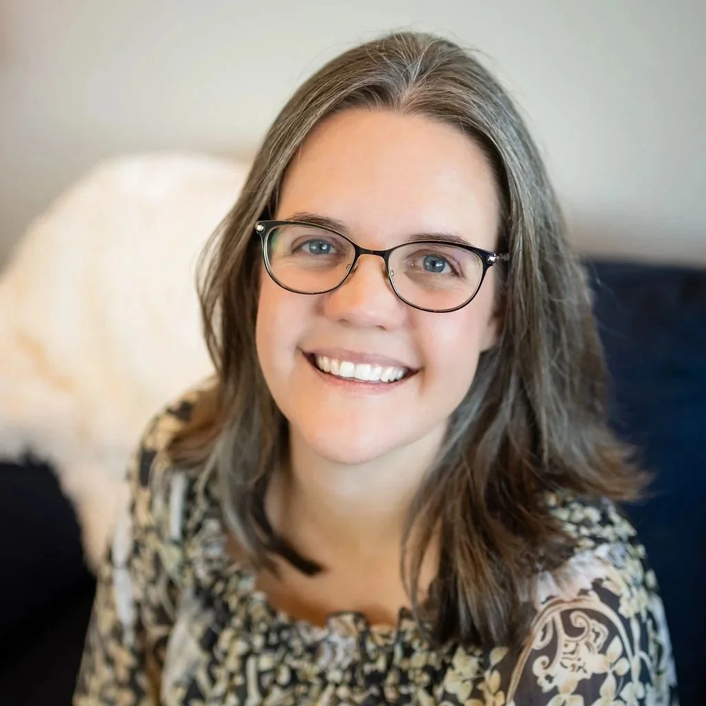 A woman with glasses and long brown hair smiling at the camera, standing in front of a teal wall with a decorative hanging textile art in the background.