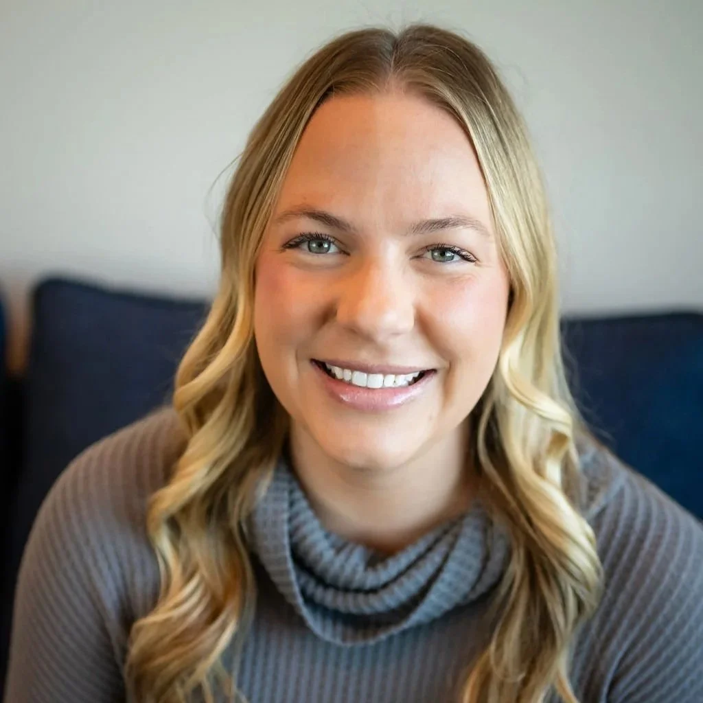 A young woman with long blonde hair smiling, wearing a dark gray T-shirt and black pants, standing in front of a blue wall with a decorative hanging made of white strings.