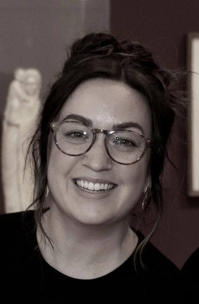 A smiling woman with glasses, dark hair, and a nose piercing, in a dimly lit indoor setting.