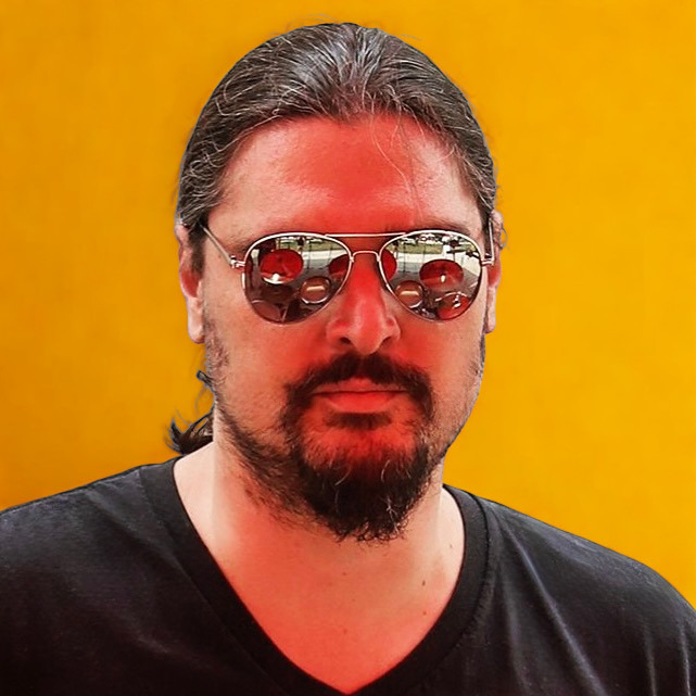 A man with dark hair, beard, and mustache wearing sunglasses with red-tinted lenses and a black shirt, standing against a yellow background.