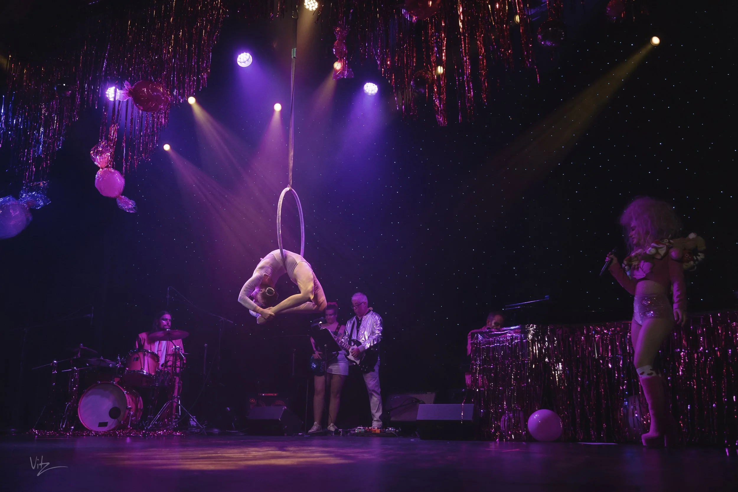 A stage with pink, purple, and gold decorations including balloons and tinsel, featuring a performer in a pink leotard hanging from a hoop, with live band and a person singing into a microphone.