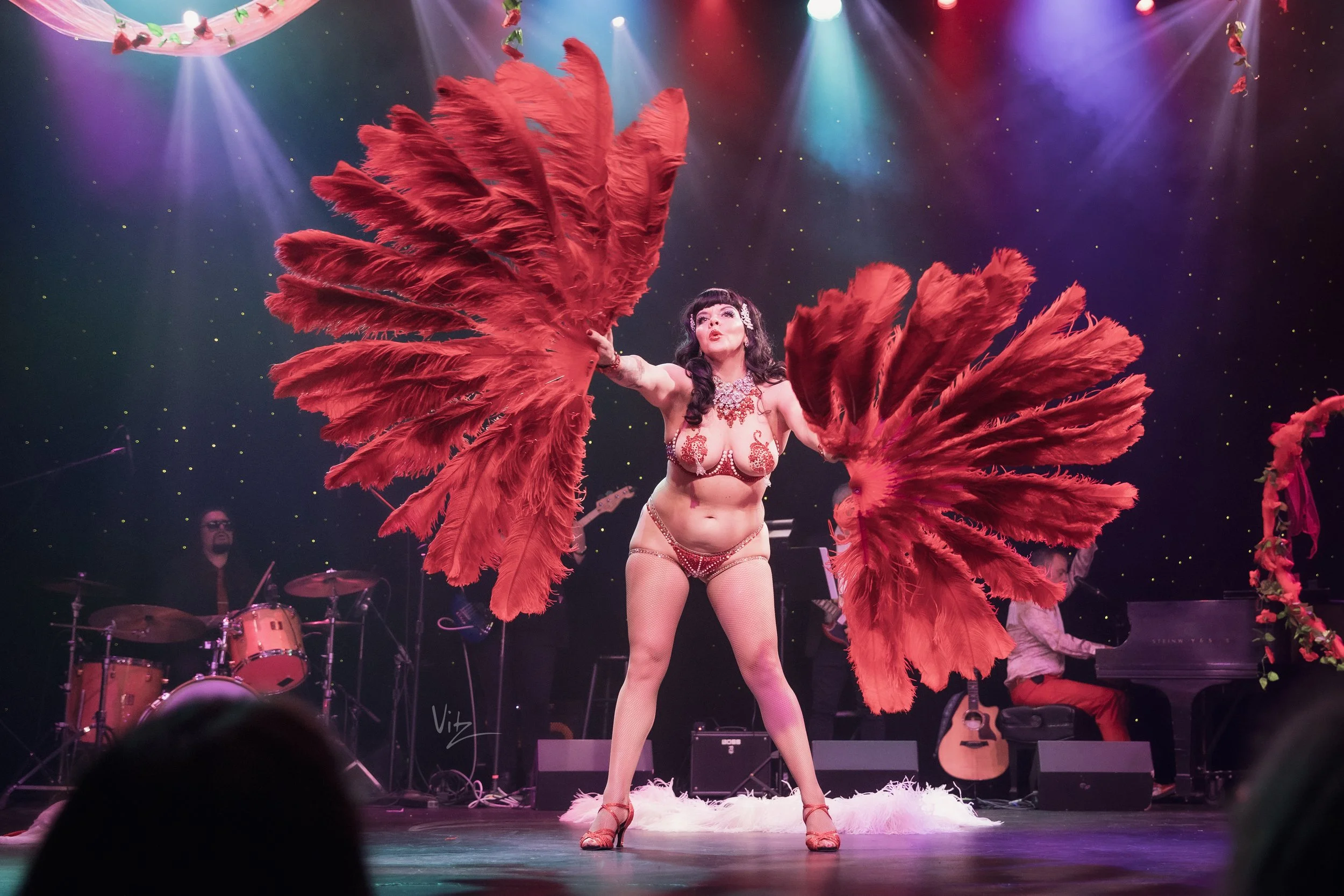 A woman dressed in a burlesque costume with large red feathered wings on stage. She has dark hair with a hair accessory, and is wearing a revealing outfit with jewelry. Stage lights shine down on her with a dark backdrop dotted with small lights. Mus