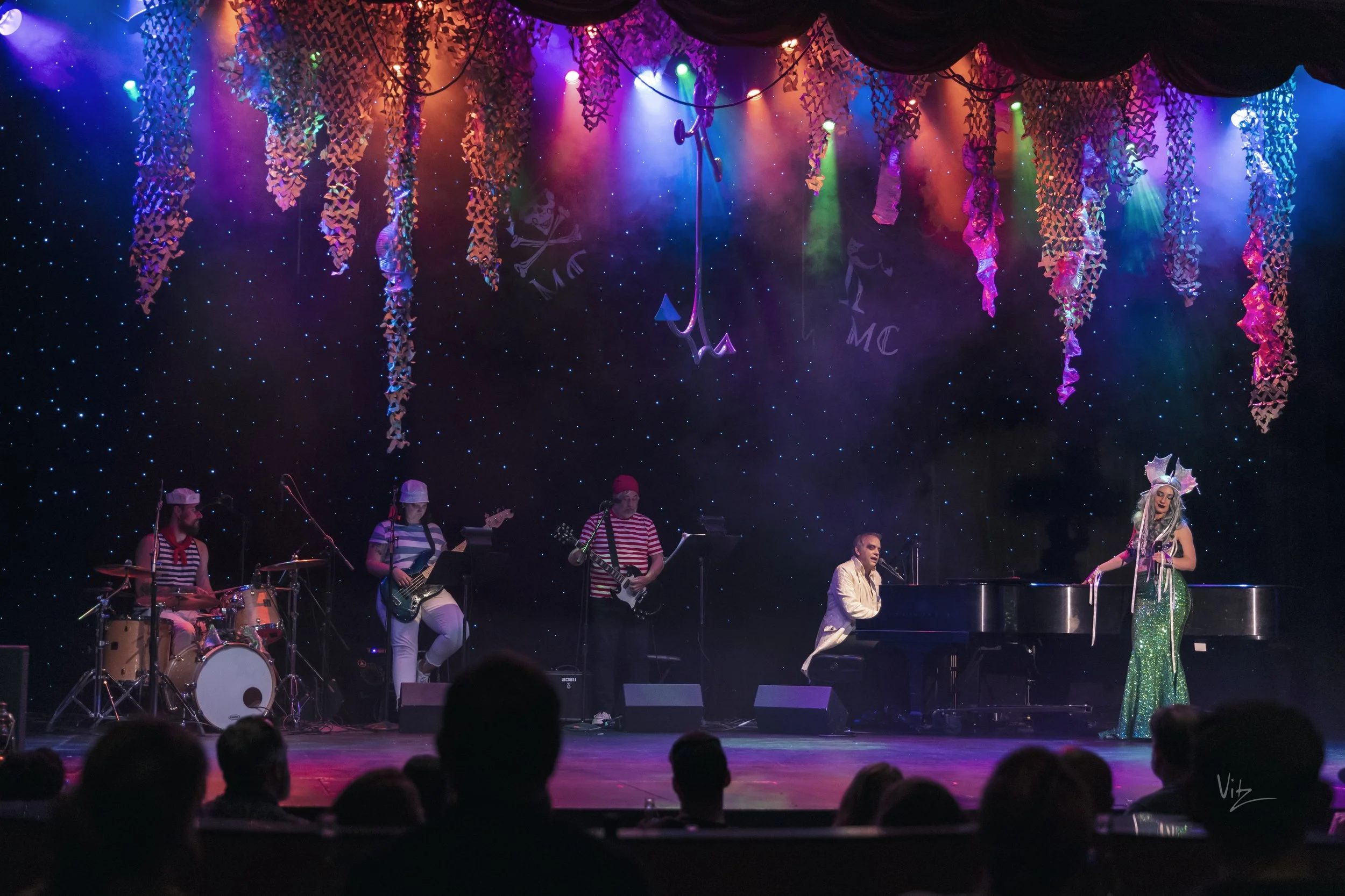 A musical stage performance with musicians dressed in colorful costumes, including striped shirts, and a performer in a sequined dress and elaborate headpiece. The stage is decorated with hanging paper chains, colorful lights, and a backdrop featurin