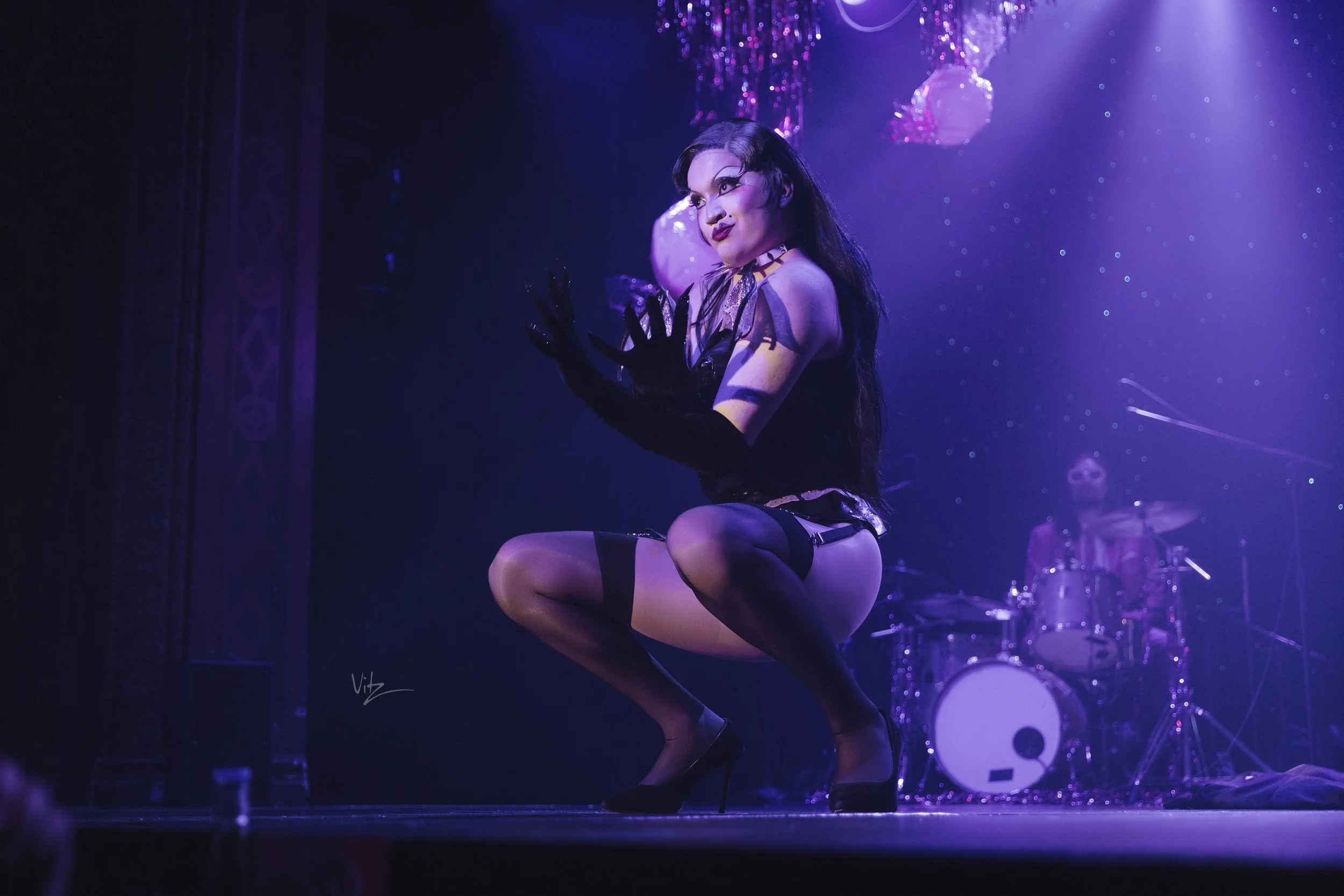 A woman performing a dance on stage, dressed in black lingerie and stockings, with purple lighting and a drum set in the background.