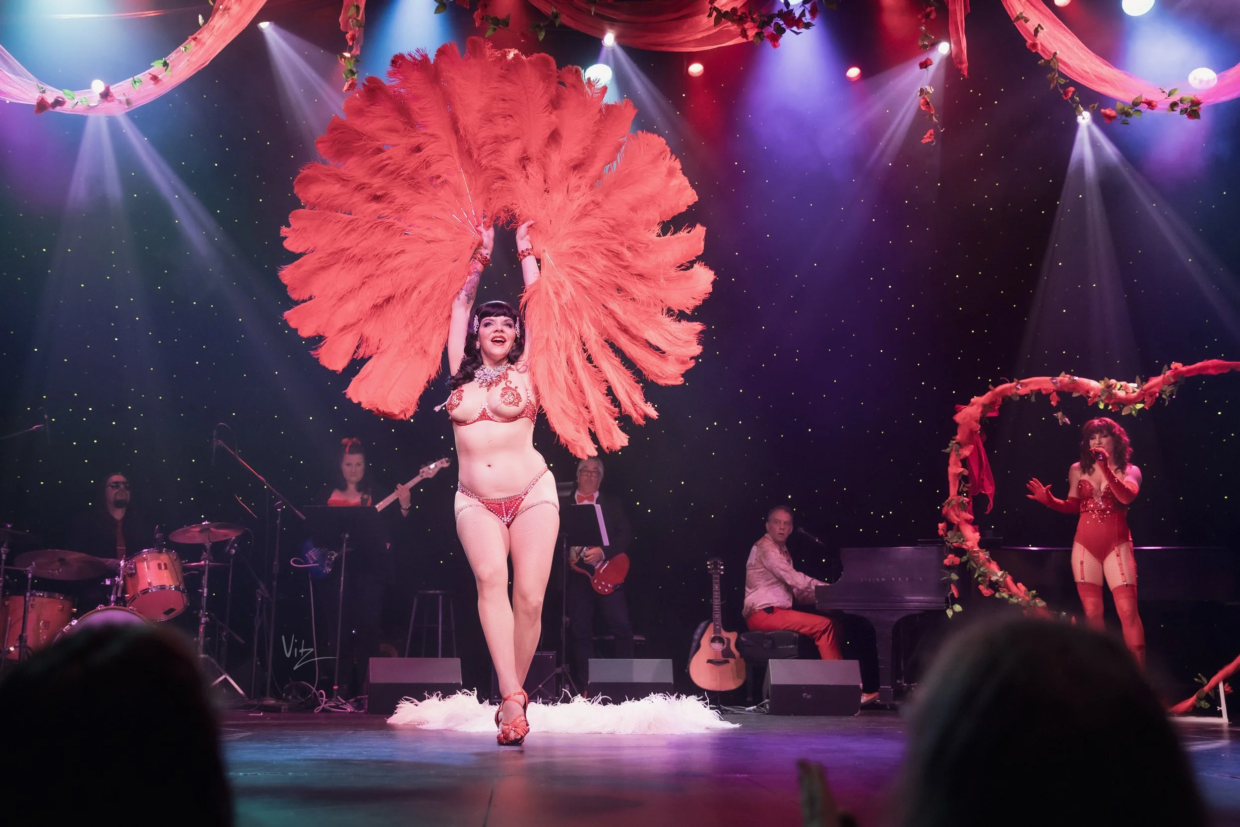 A performer on stage dressed in a revealing costume with large red feathers, smiling, with musicians and stage lights in the background.