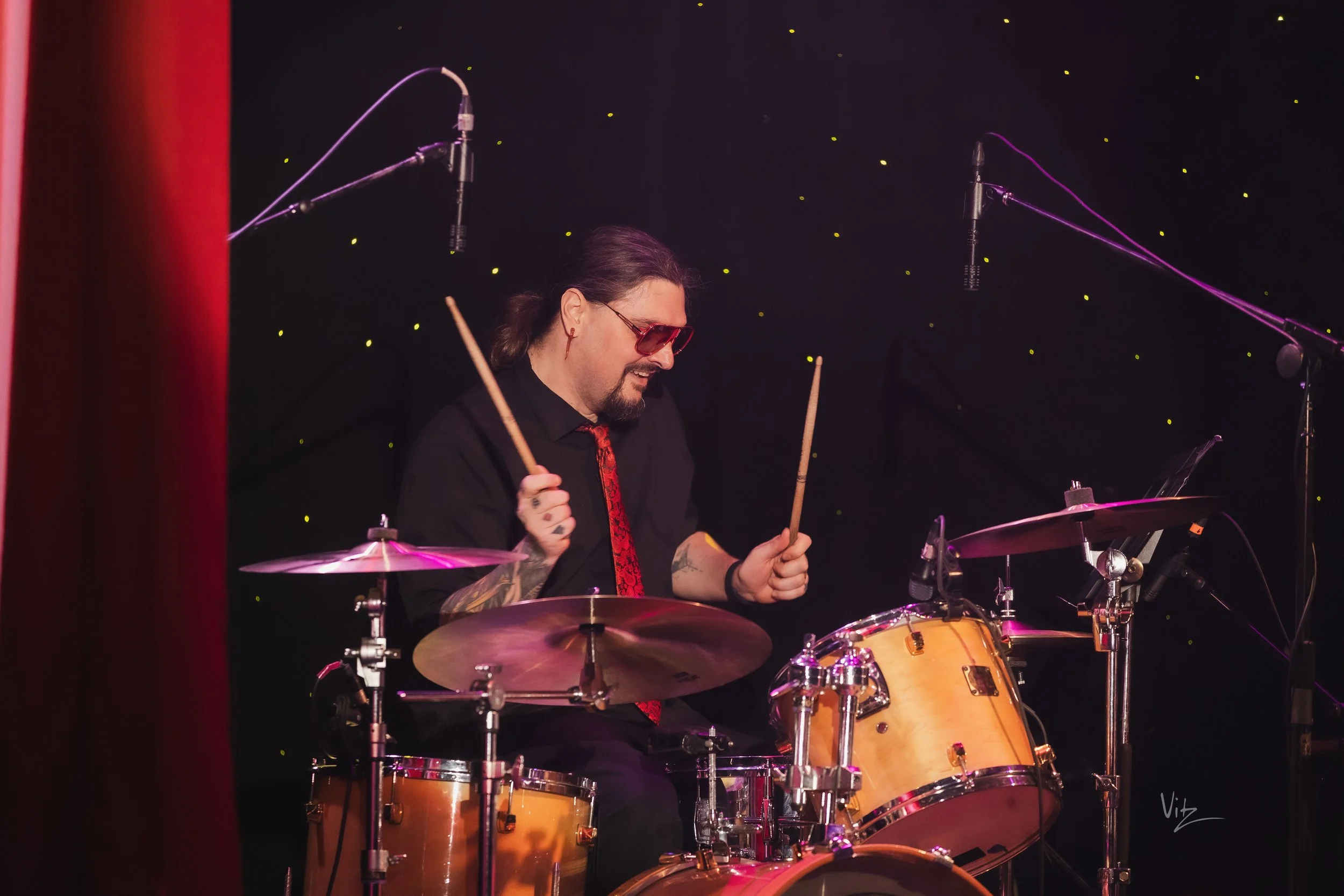 A man with long hair, sunglasses, and tattooed arms playing a drum set with a dark background featuring small yellow lights and microphones overhead.