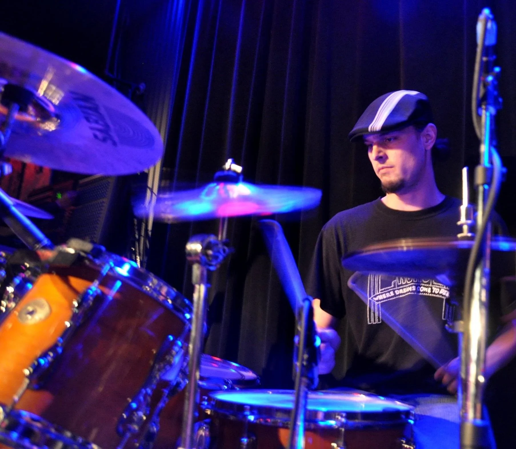 A musician wearing a black t-shirt and a hat with parallel white stripes plays drums on stage under blue lighting.