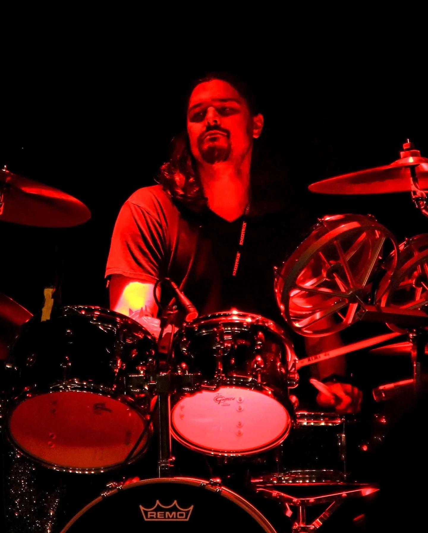 A man with long hair, beard, and glasses playing a drum set illuminated by red lighting at night.