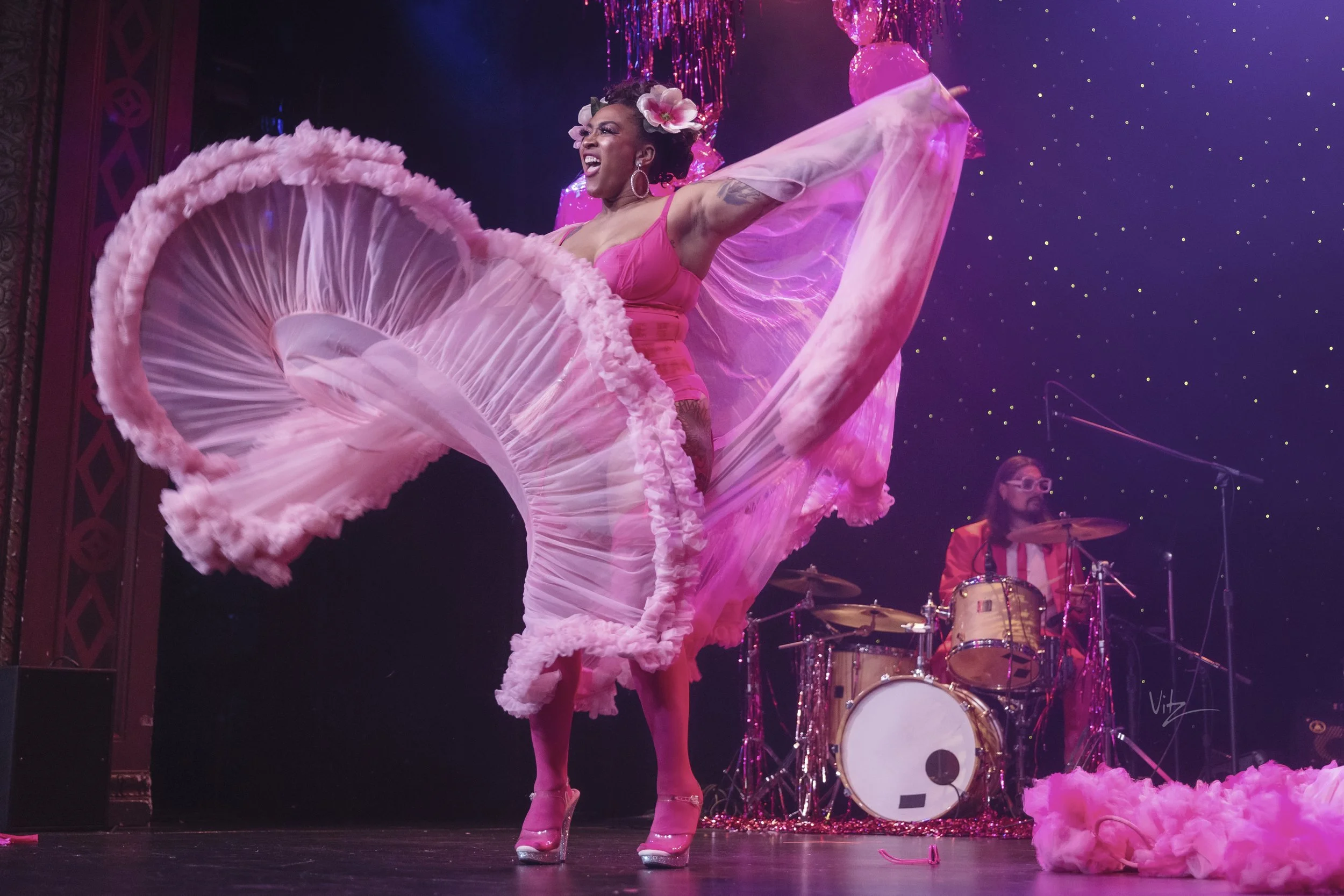 A woman in a pink dress and high heels dancing on stage with her arms raised, surrounded by pink fabric and flowers, with a band playing in the background under purple and pink lights.