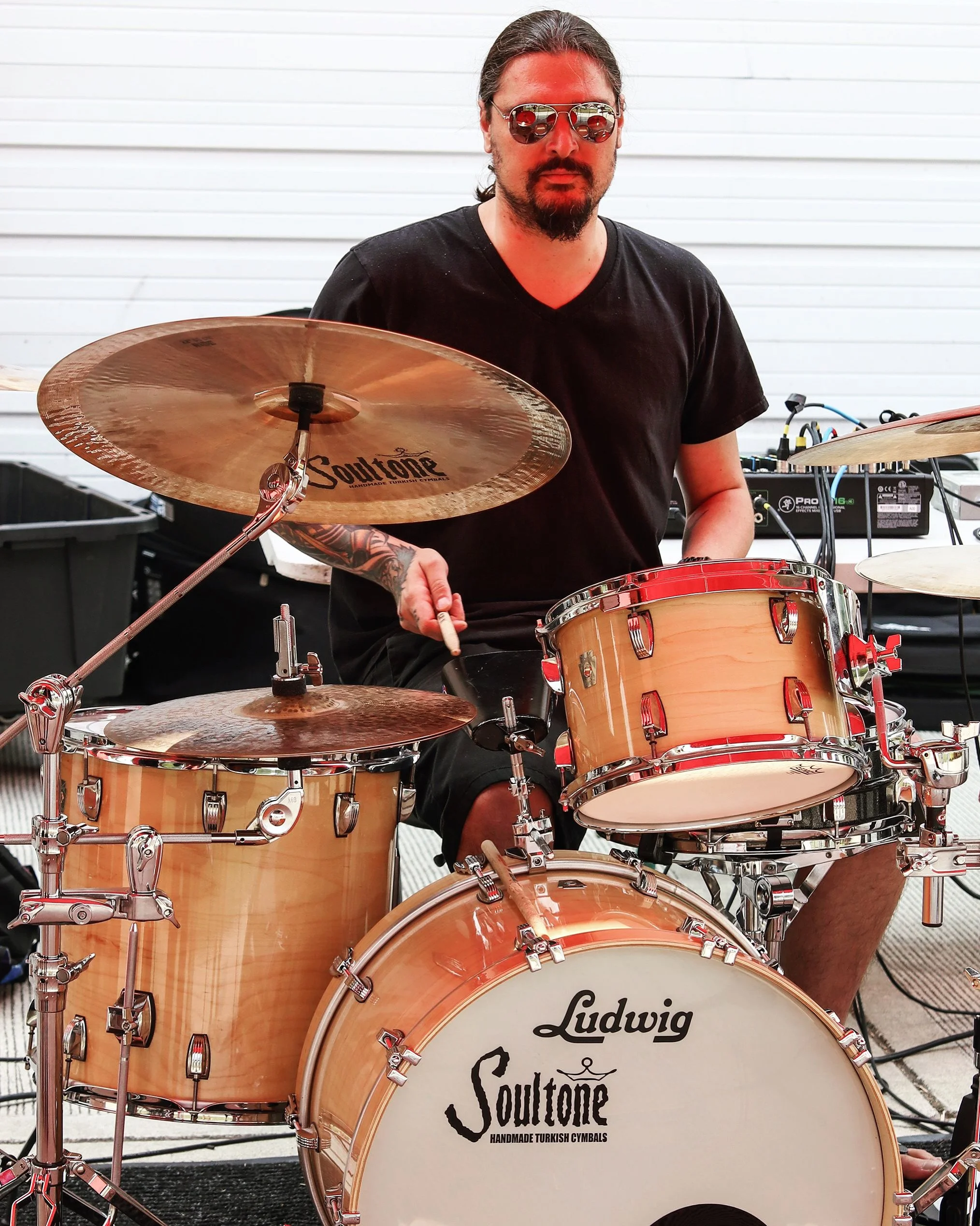 A man with long hair and sunglasses playing a drum set outdoors. The drum set has a logo that reads "SoulTone" and "Handmade Turkish Cymbals." The man has a tattoo on his left forearm and is wearing a black t-shirt.