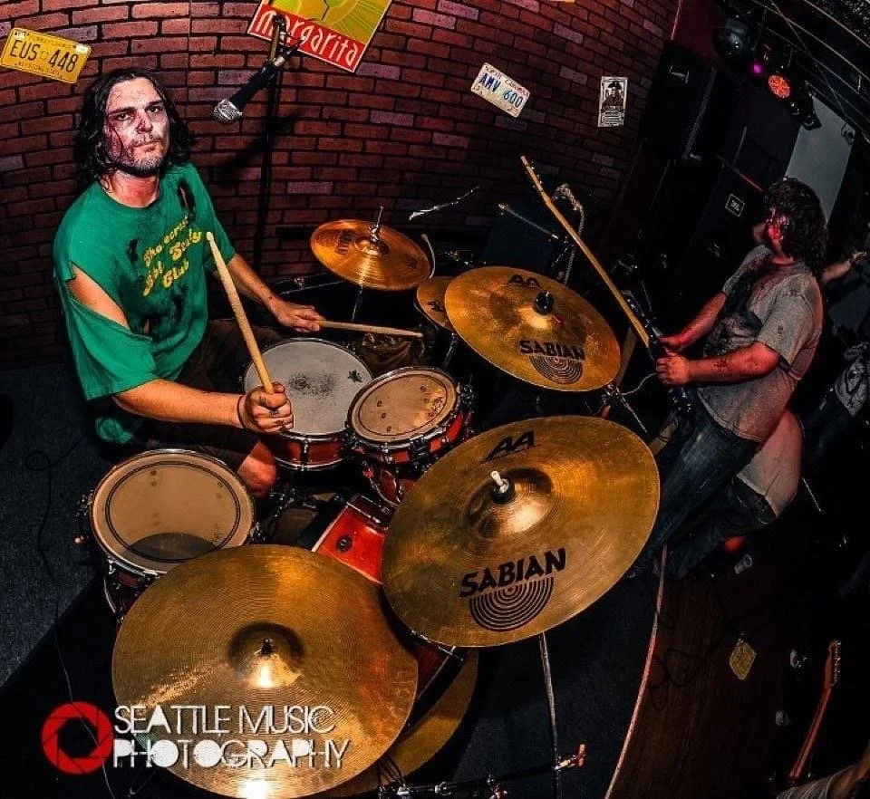 A drummer playing a drum set with cymbals labeled Sabian, in a brick-walled music venue. The drummer has long dark hair and is wearing a green shirt, holding drumsticks. There is another musician in the background with long curly hair and tattoos, ho