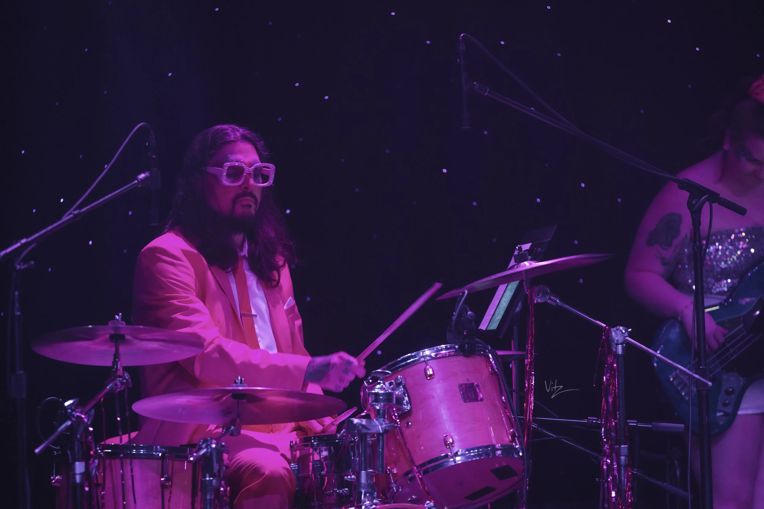 A musician in a pink suit and large glasses playing a drum set on a dark stage with purple lighting.