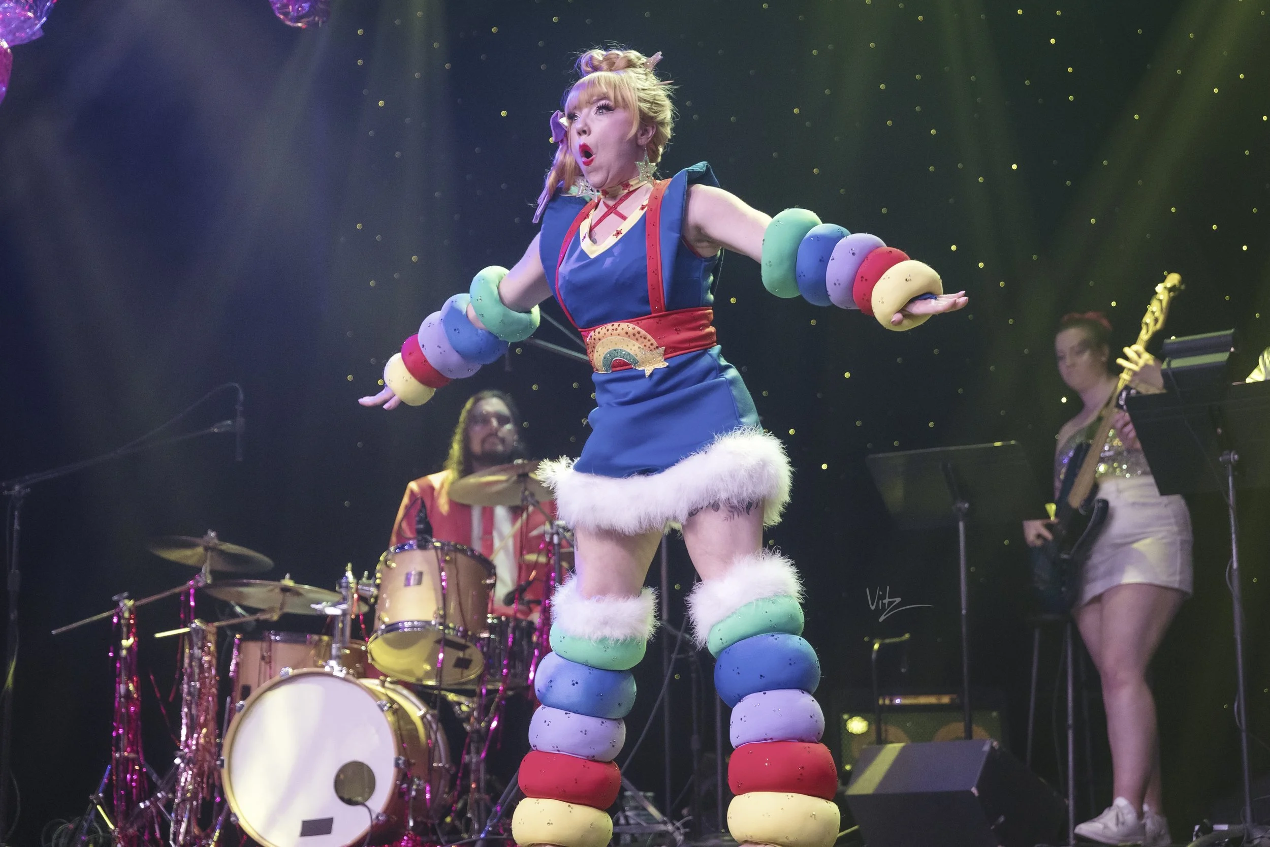 A woman with red hair styled in a bun, wearing a colorful and whimsical costume, stands on stage with her arms outstretched, surrounded by musicians. She has rainbow-colored arm and leg warmers made of stacked bouncy balls and white fluffy trim at he
