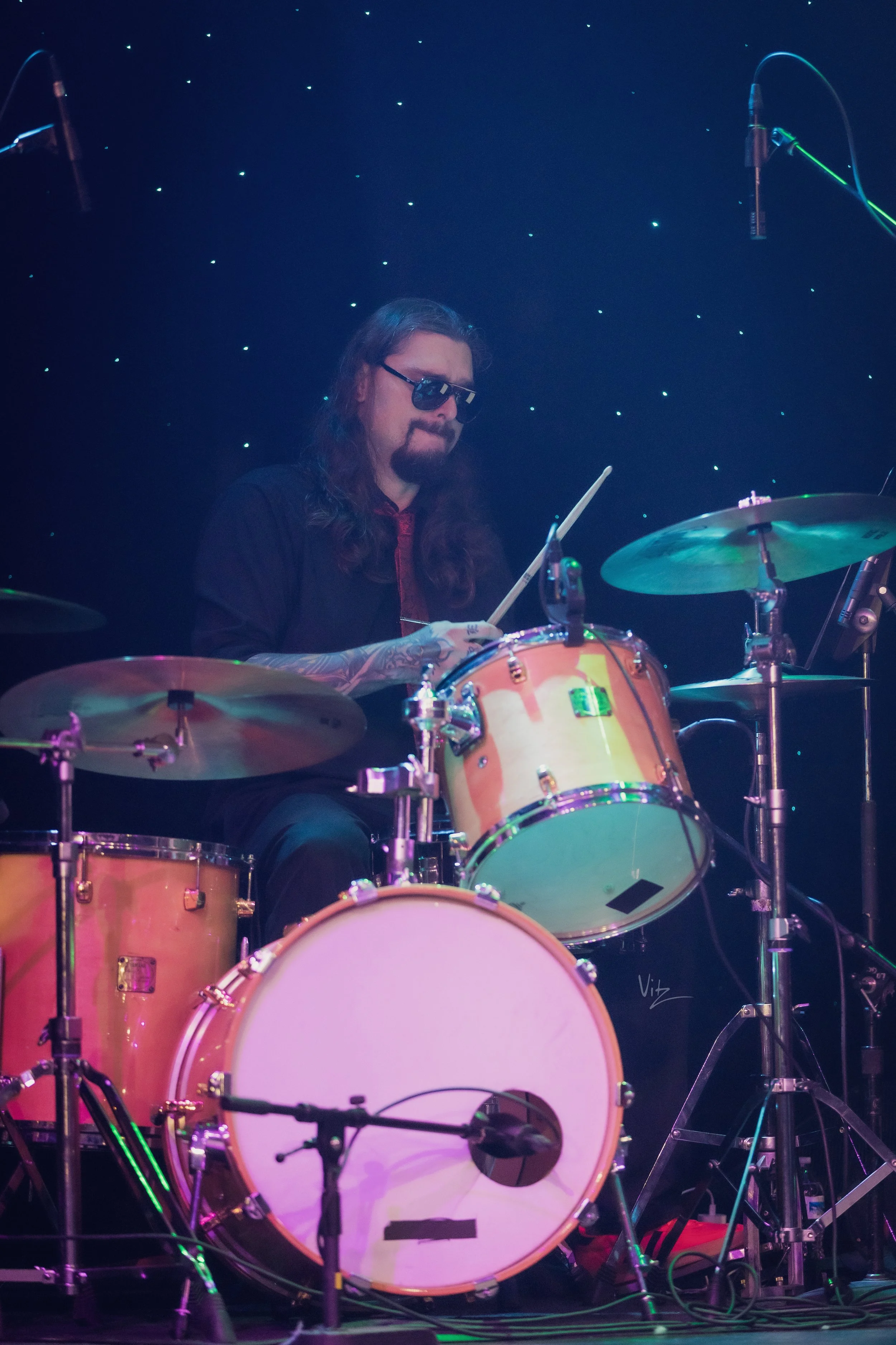 A man with long hair, wearing sunglasses, is playing a drum set on a stage with a starry backdrop.