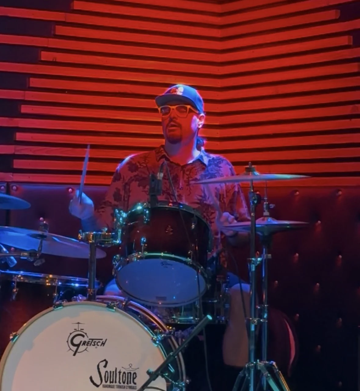 A man wearing sunglasses and a cap playing a Gretsch drum set against a red-lit wooden wall background.