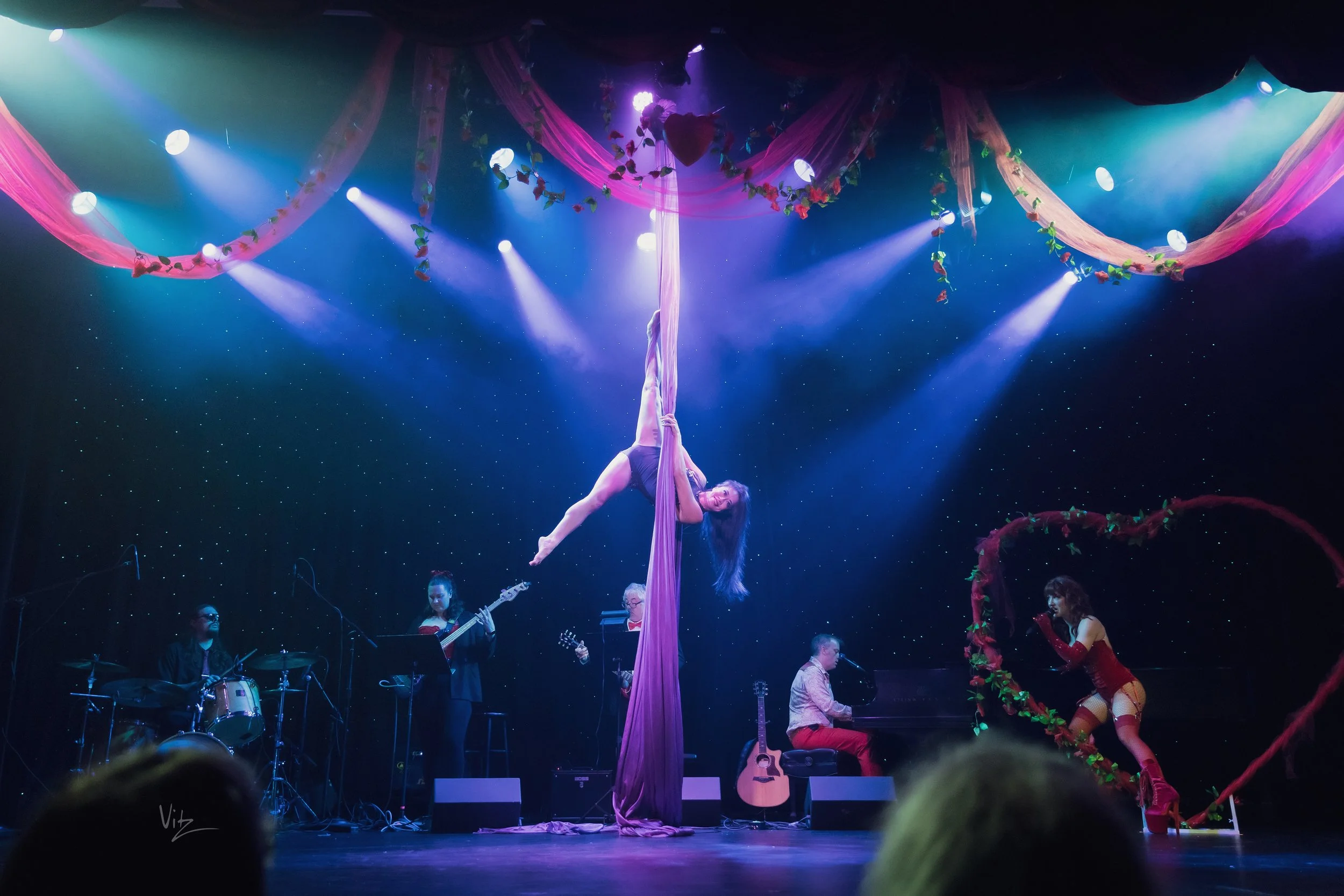A stage performance with a woman performing an aerial silk act, surrounded by live musicians and backup dancers, with colorful lighting and decorations.