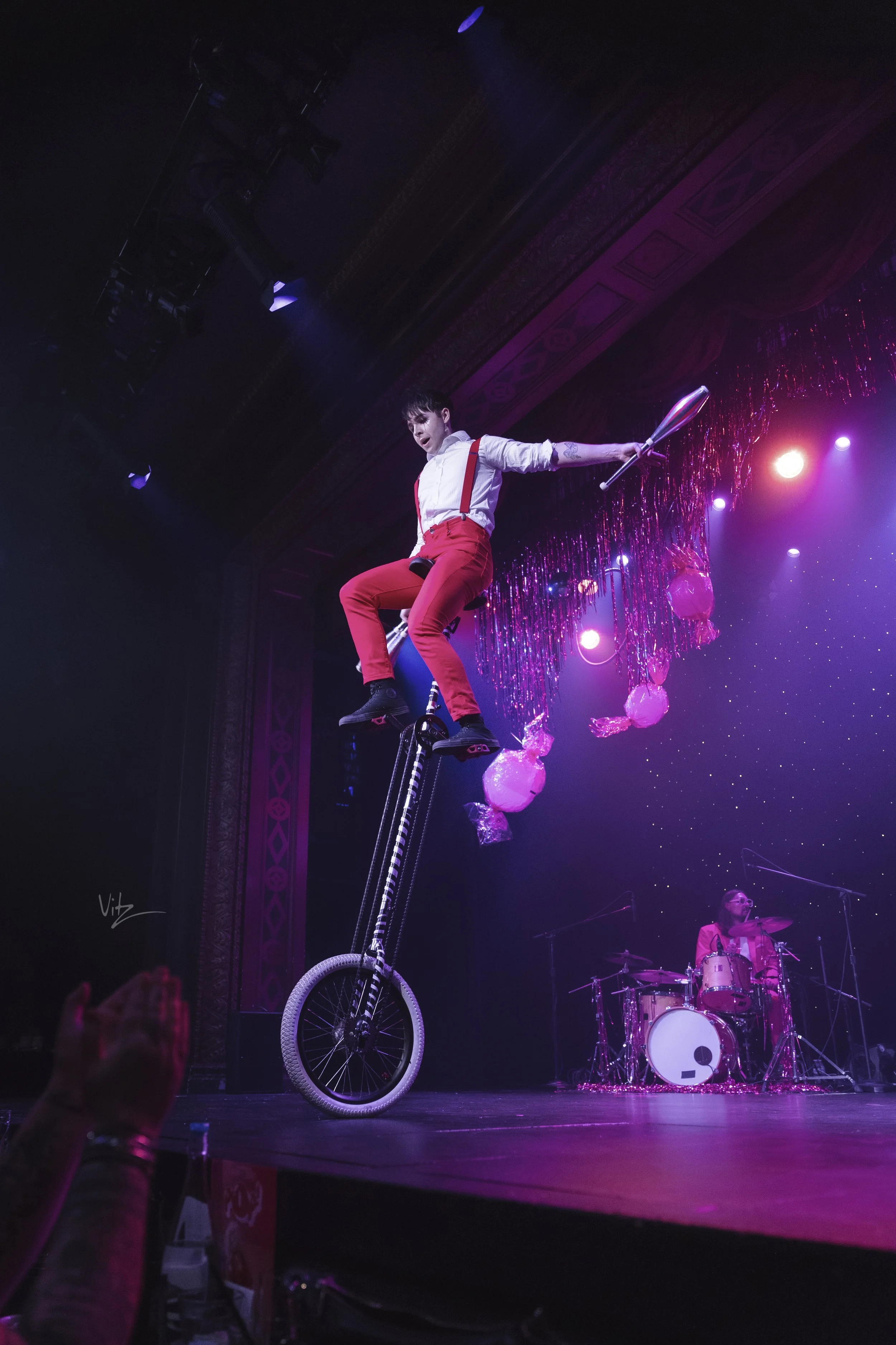 Performer dressed in white shirt with red suspenders and red pants on a unicycle during a stage performance, with colorful stage lights and a drummer in the background.