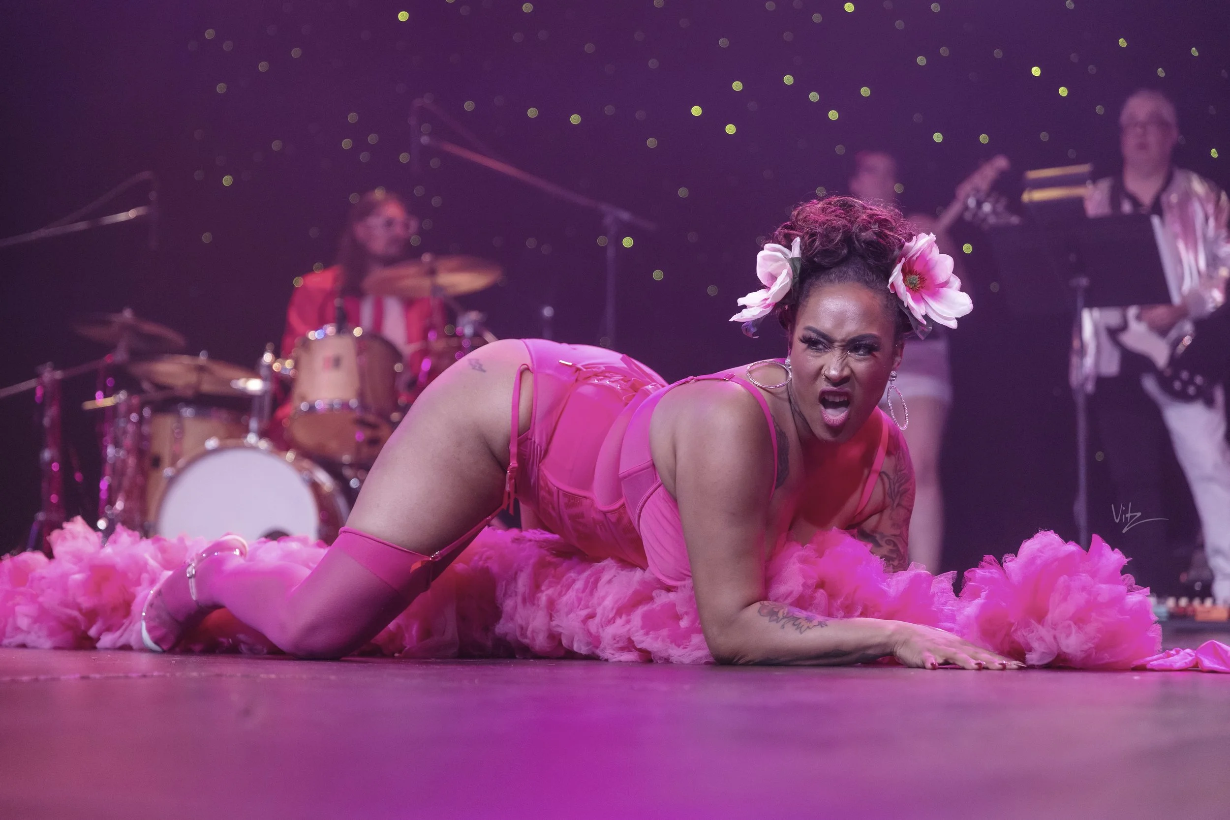 A performer with dark hair styled in an updo, wearing pink lingerie, thigh-high stockings, and flowers in her hair, on stage surrounded by pink feathers, with a band playing in the background against a starry backdrop.