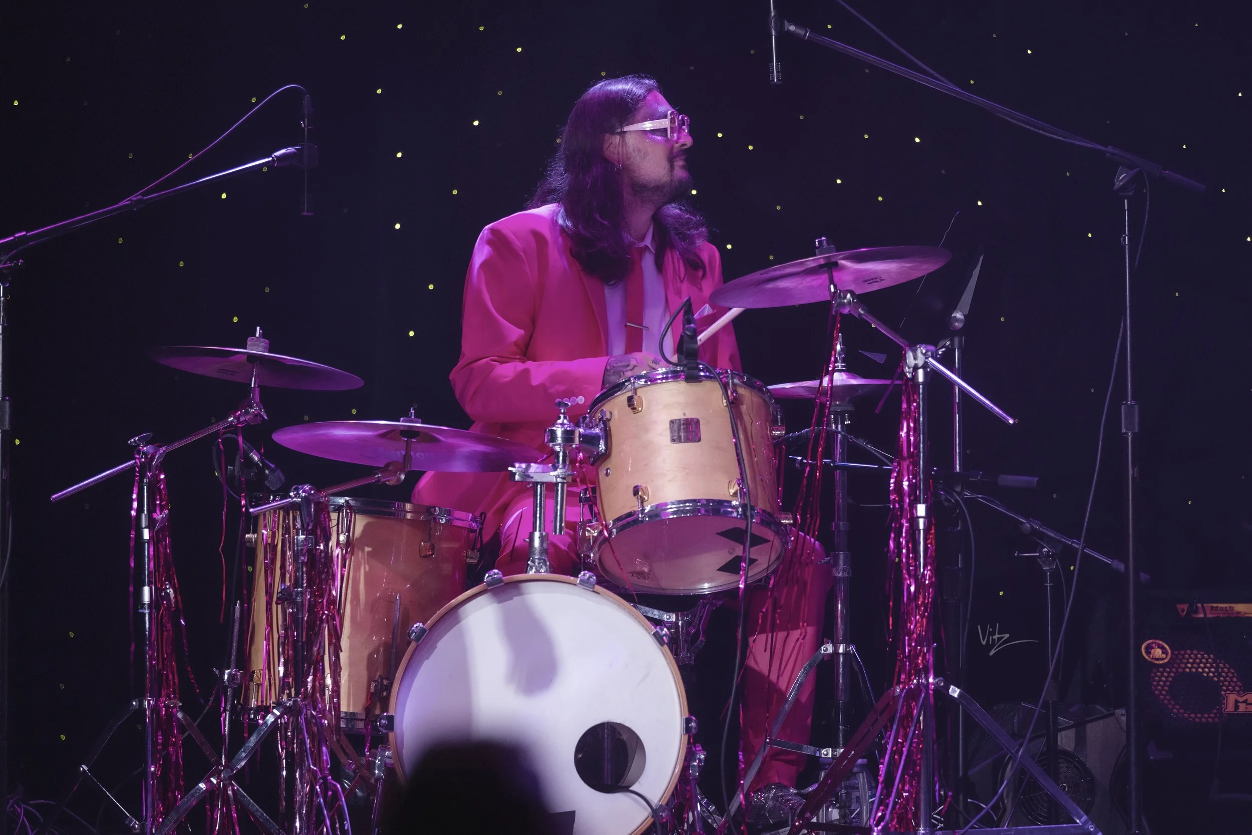 A man with long hair and glasses wearing a pink suit playing a drum set on stage under purple lighting and starry background.