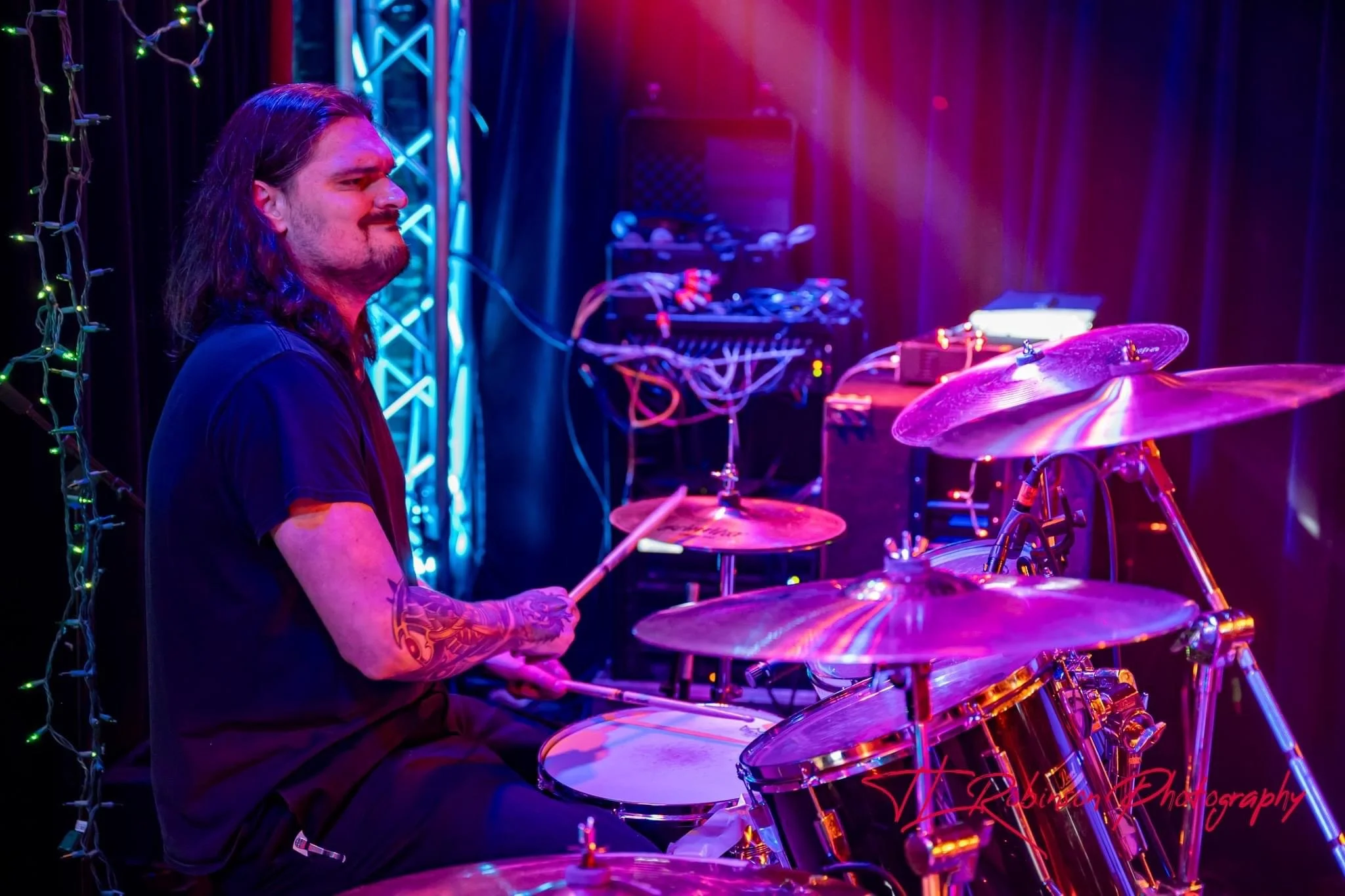 A man with long hair and tattoos playing the drums on stage, illuminated by purple, pink, and blue stage lights with decorative string lights and audio equipment in the background.