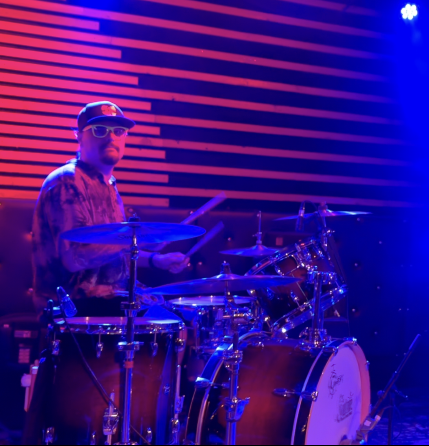 A drummer playing a drum set under colorful stage lighting with pink and blue hues, background with horizontal neon lights.