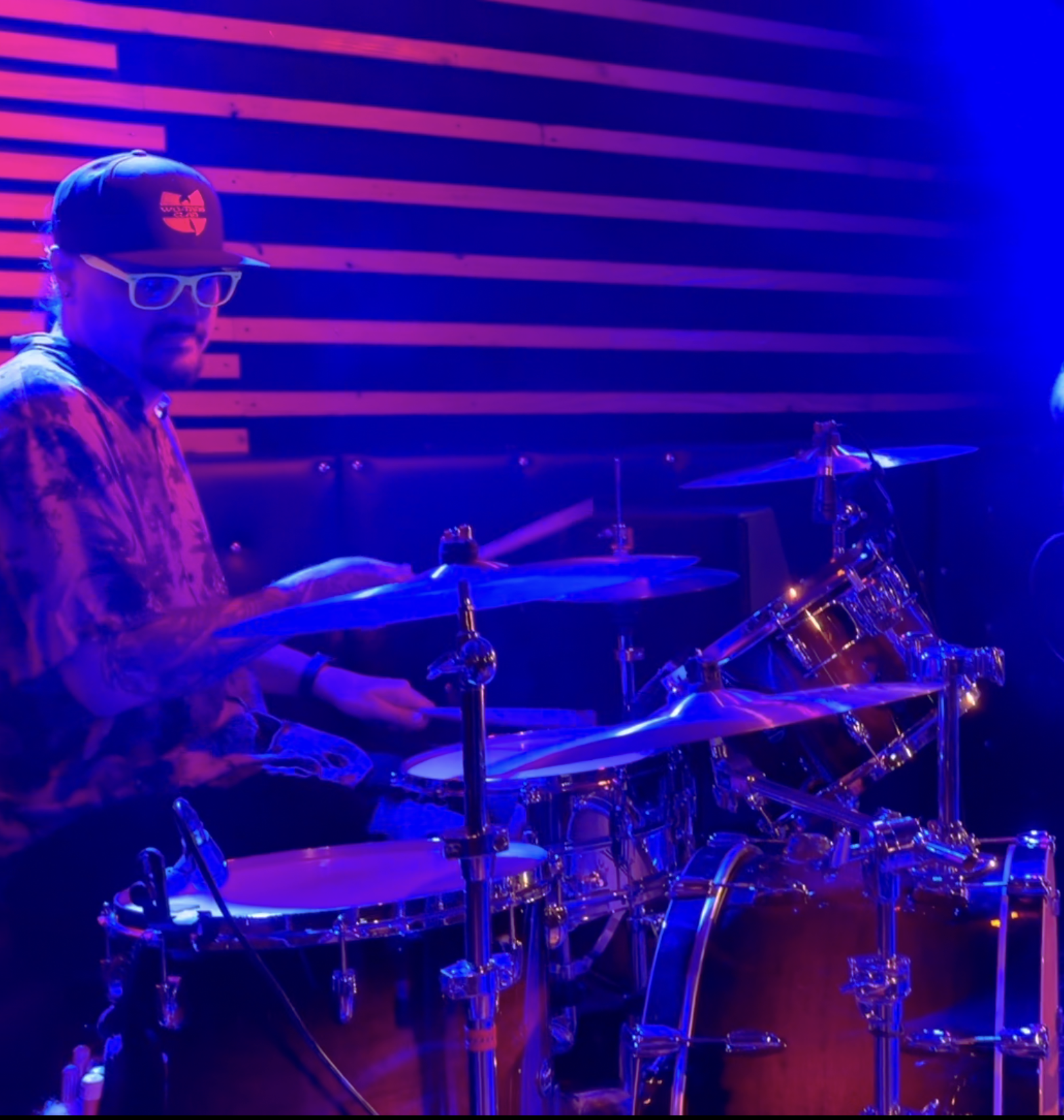 A person with a beard, wearing a cap, glasses, a patterned shirt, and gloves, sitting behind a drum set illuminated with purple and blue lighting in a music studio with horizontal wood paneling on the wall.