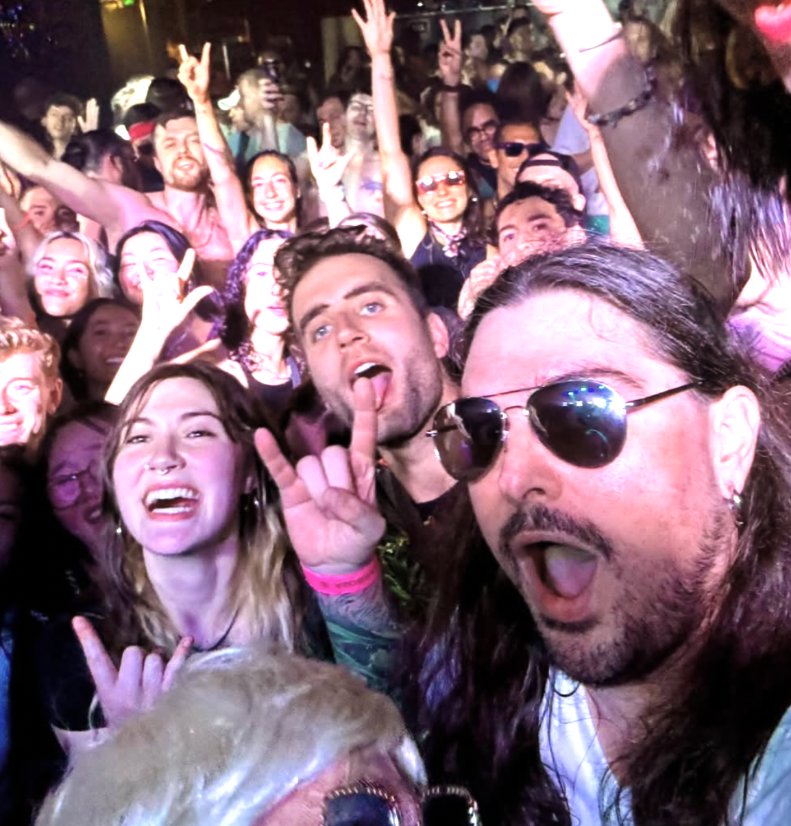A large group of people at a lively party or concert, many smiling, making peace signs, and sticking out their tongues, in a crowded indoor venue with colorful lighting.