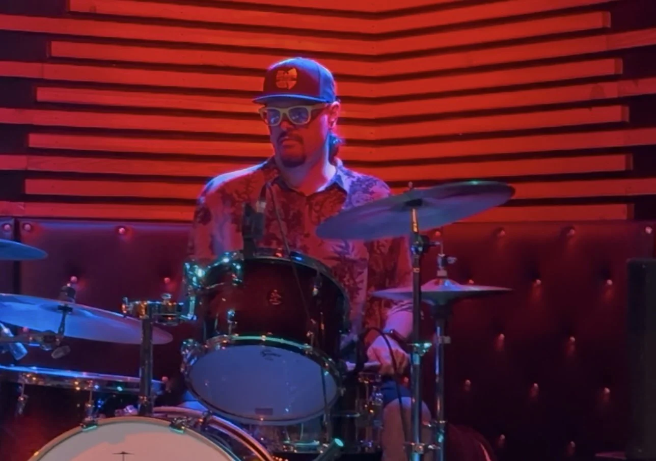 A man wearing sunglasses and a baseball cap playing a drum set in a dimly lit venue with a red and black wall background.