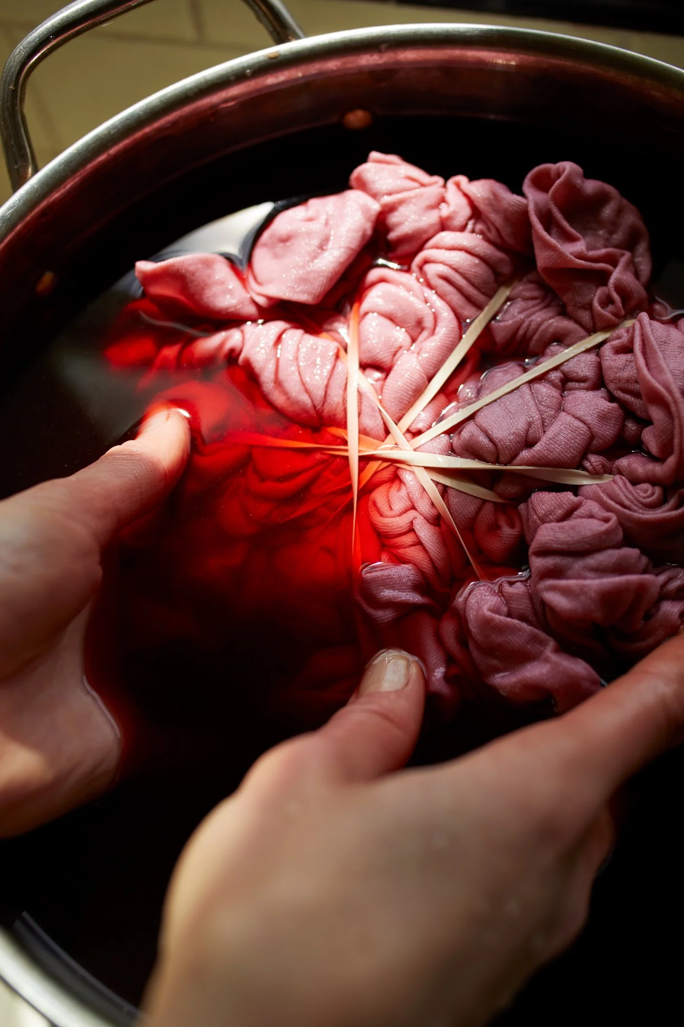 Hands holding pink fabric being dyed in red liquid with rubber bands in a pot.